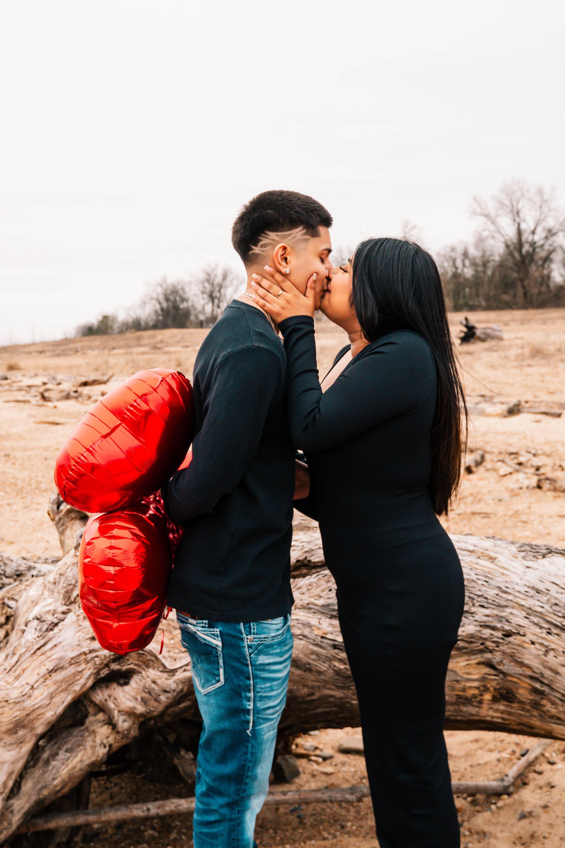 Two people kissing outdoors by a large fallen log, with one wearing a red jacket.