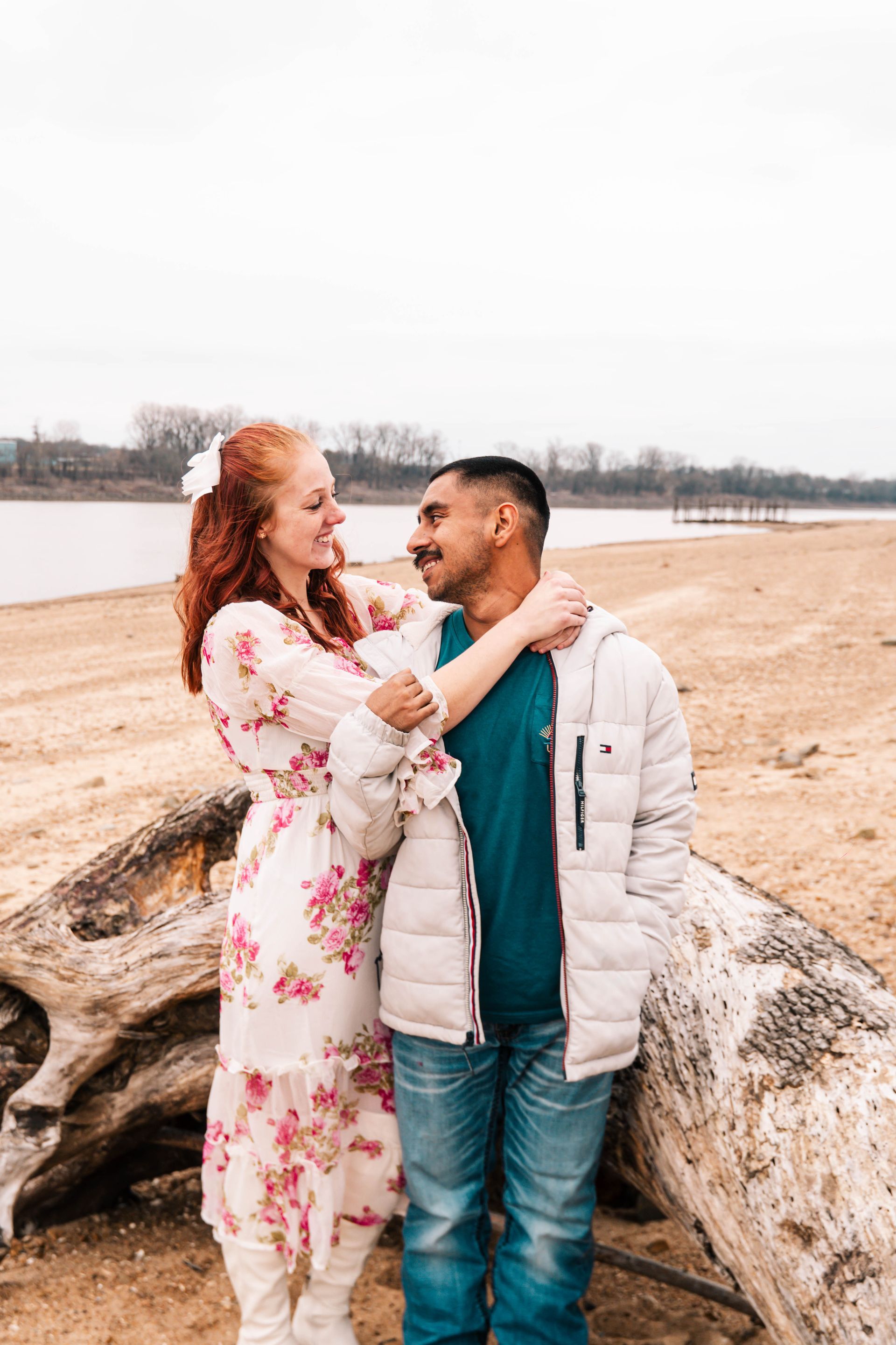 Couple smiling by a large driftwood log on a sandy beach, facing each other with water in the background