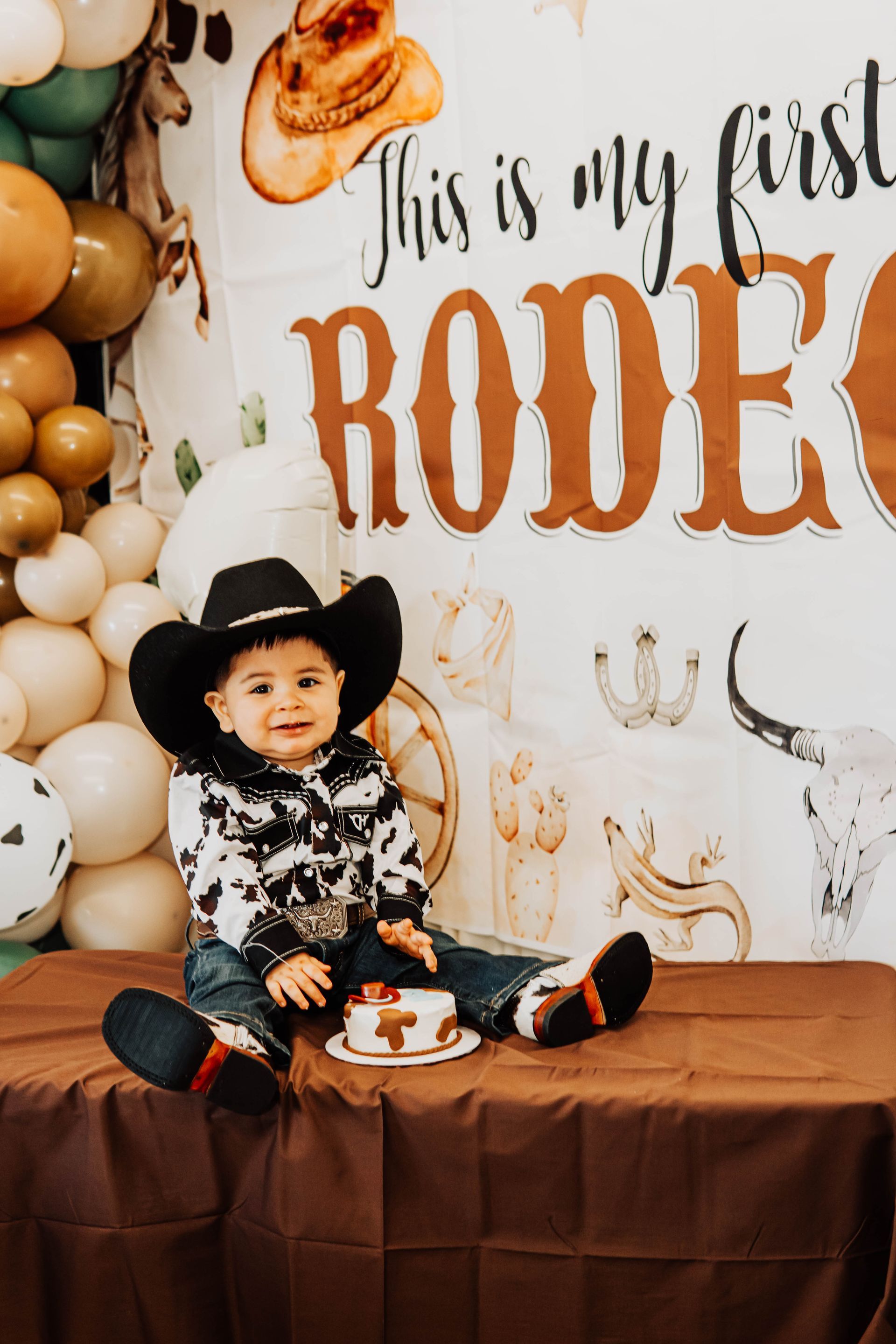 Child in cowboy hat sits at a rodeo-themed birthday setup with balloons and “This is my first rodeo” sign.