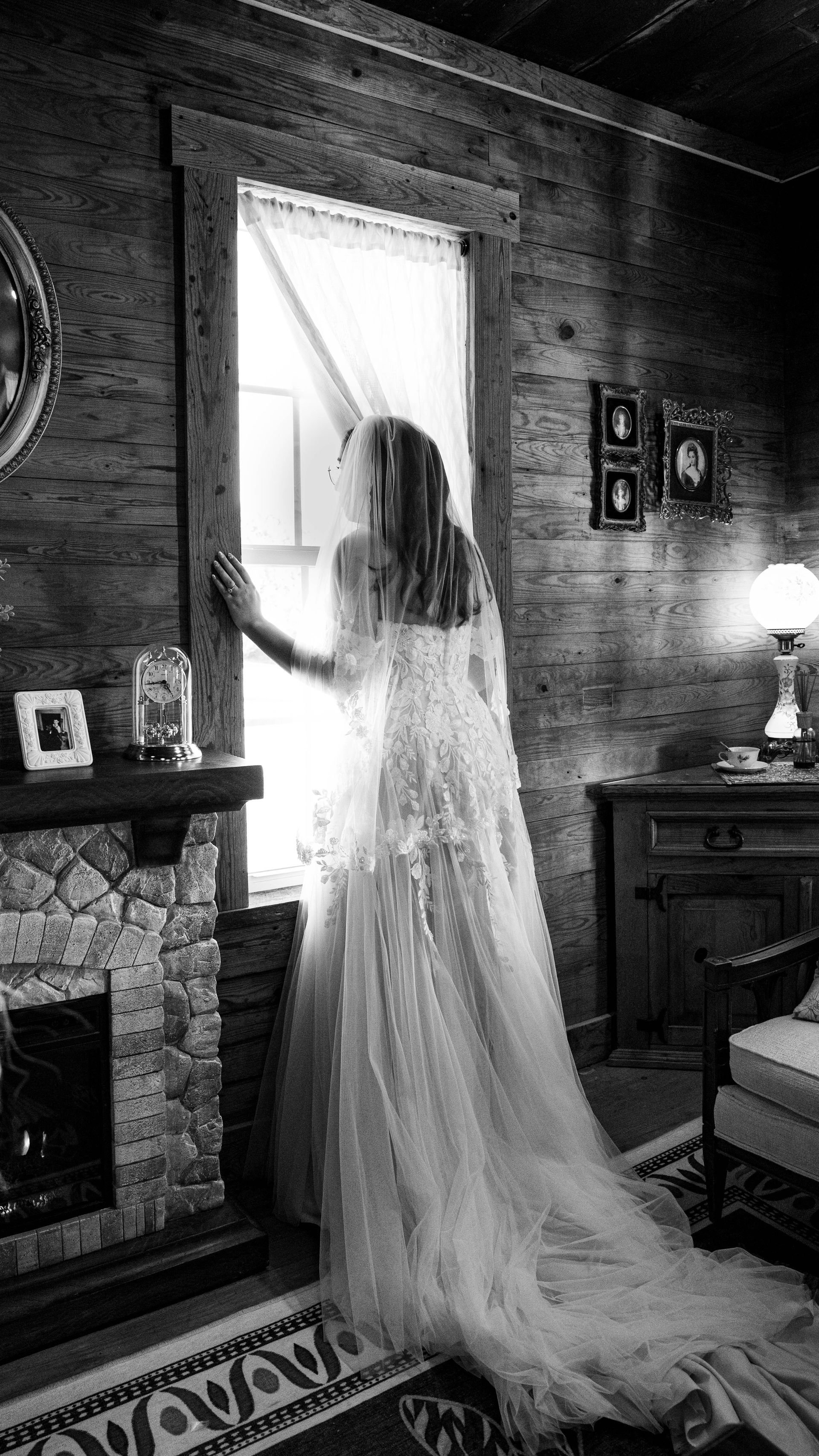 Woman in a long white gown standing by a window in a rustic, dimly lit room
