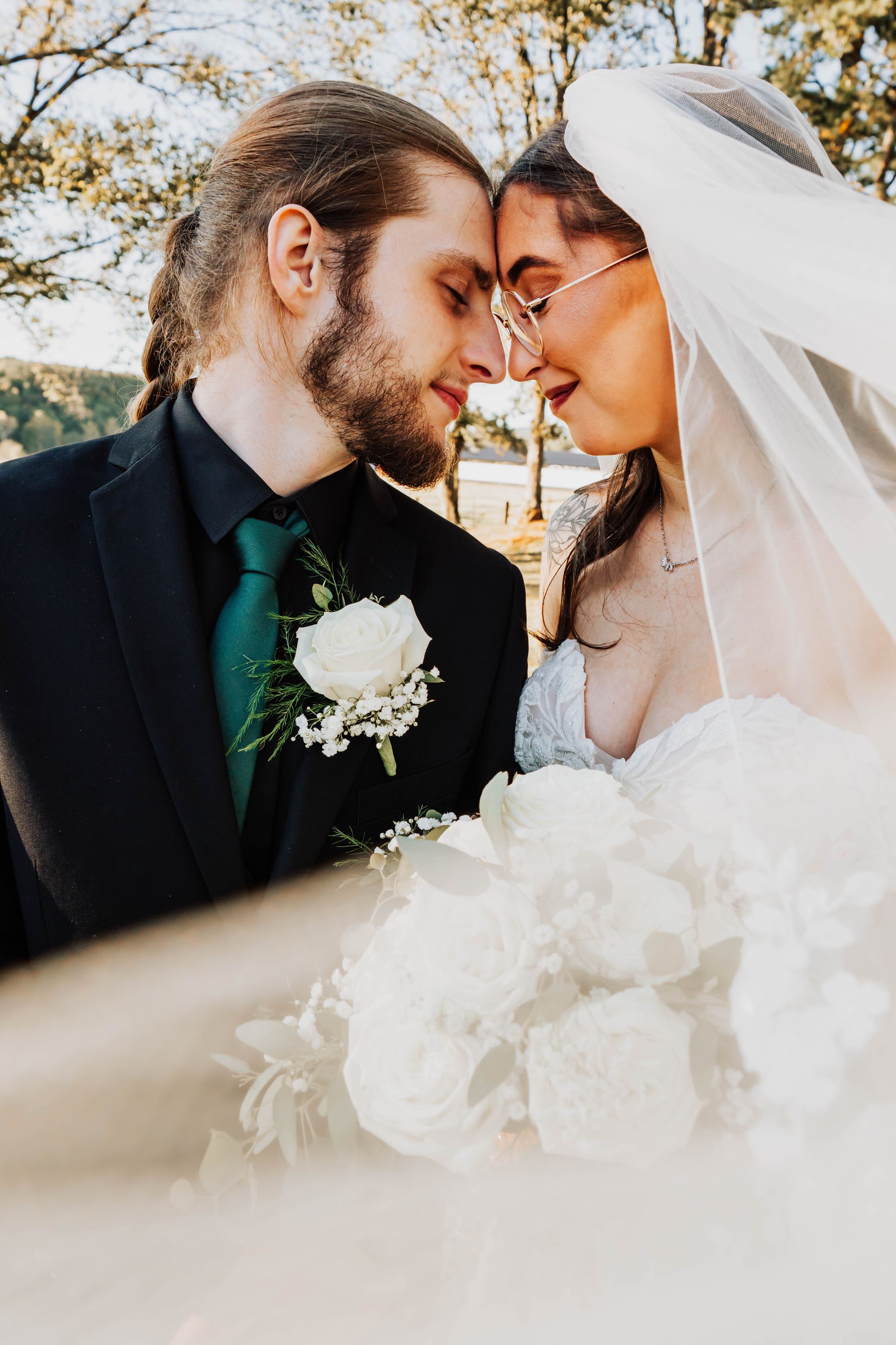 Bride and groom touch foreheads in a sunlit outdoor wedding portrait, holding white flowers.