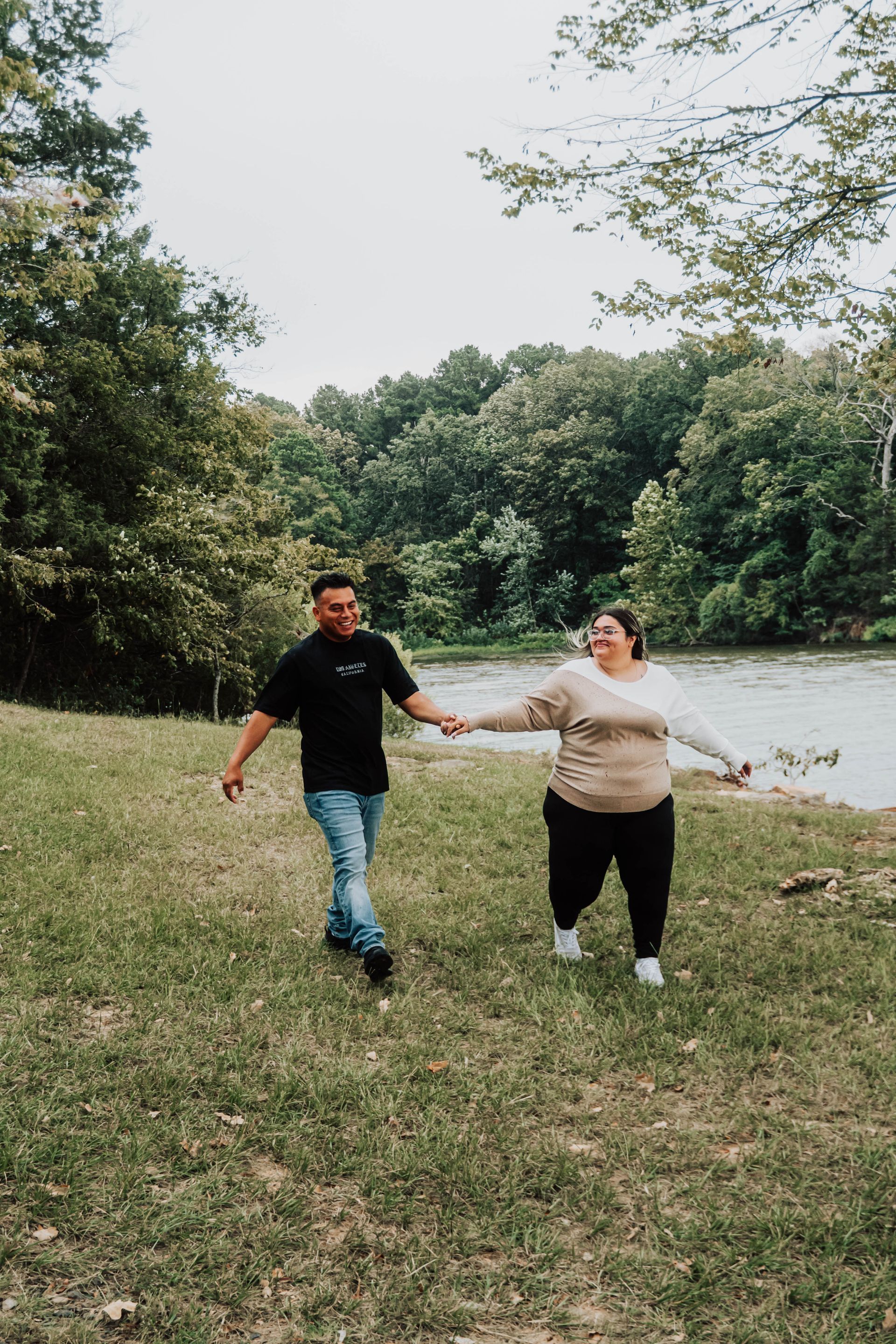 Two people holding hands and walking in a grassy park by a lake, with trees in the background.