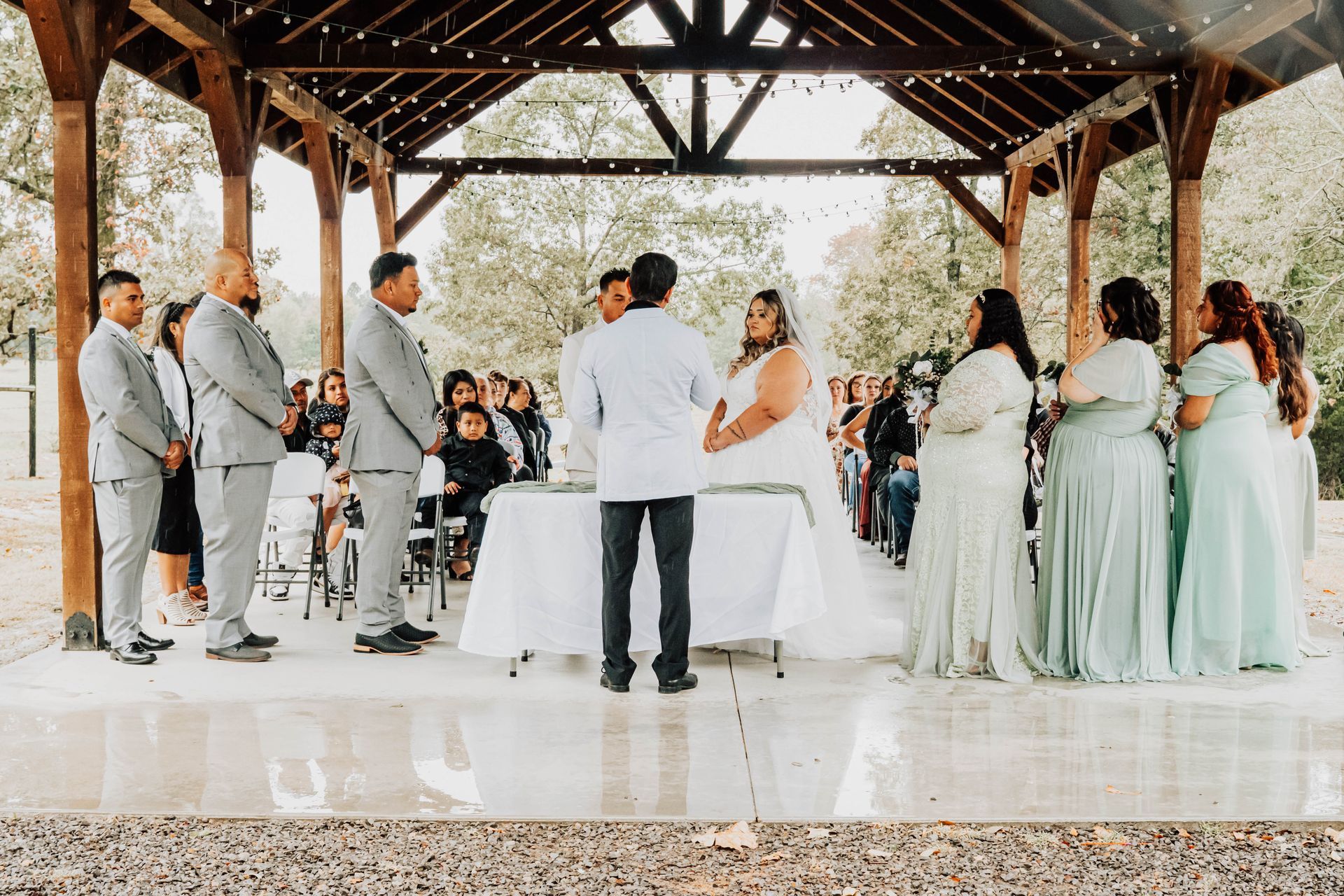 Wedding ceremony under a wooden pavilion, with the couple at the altar and guests standing nearby.