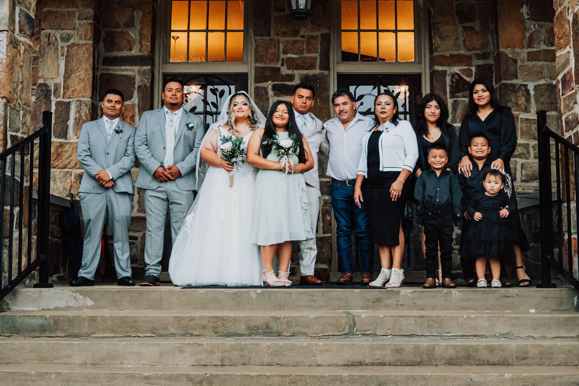 Wedding party posing on stone steps outside a building with warm-lit windows at dusk
