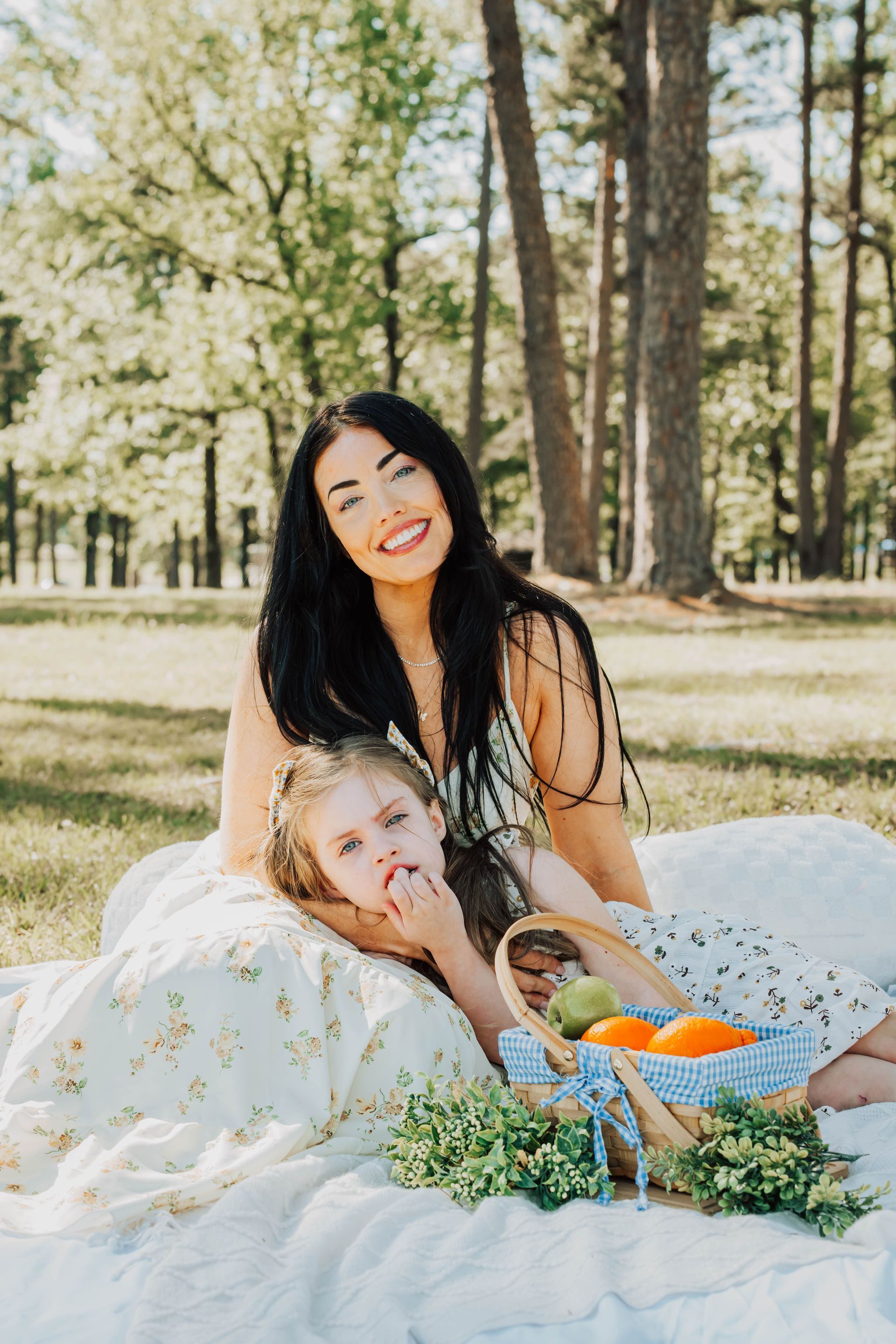 Woman and young child relaxing on a blanket in a sunny park with flowers and a basket of fruit