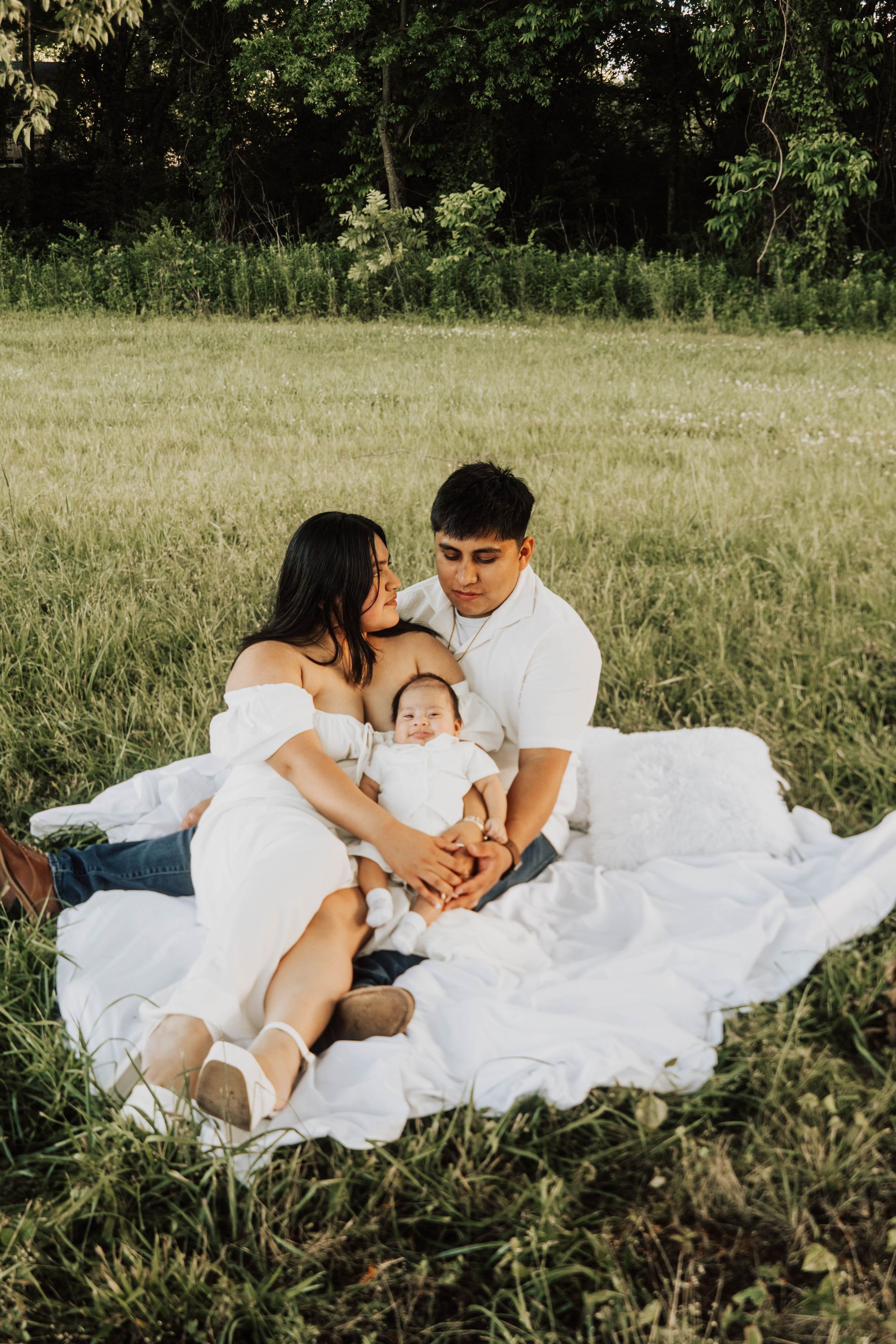 Family sitting on a white blanket in a grassy field, holding a baby outdoors.