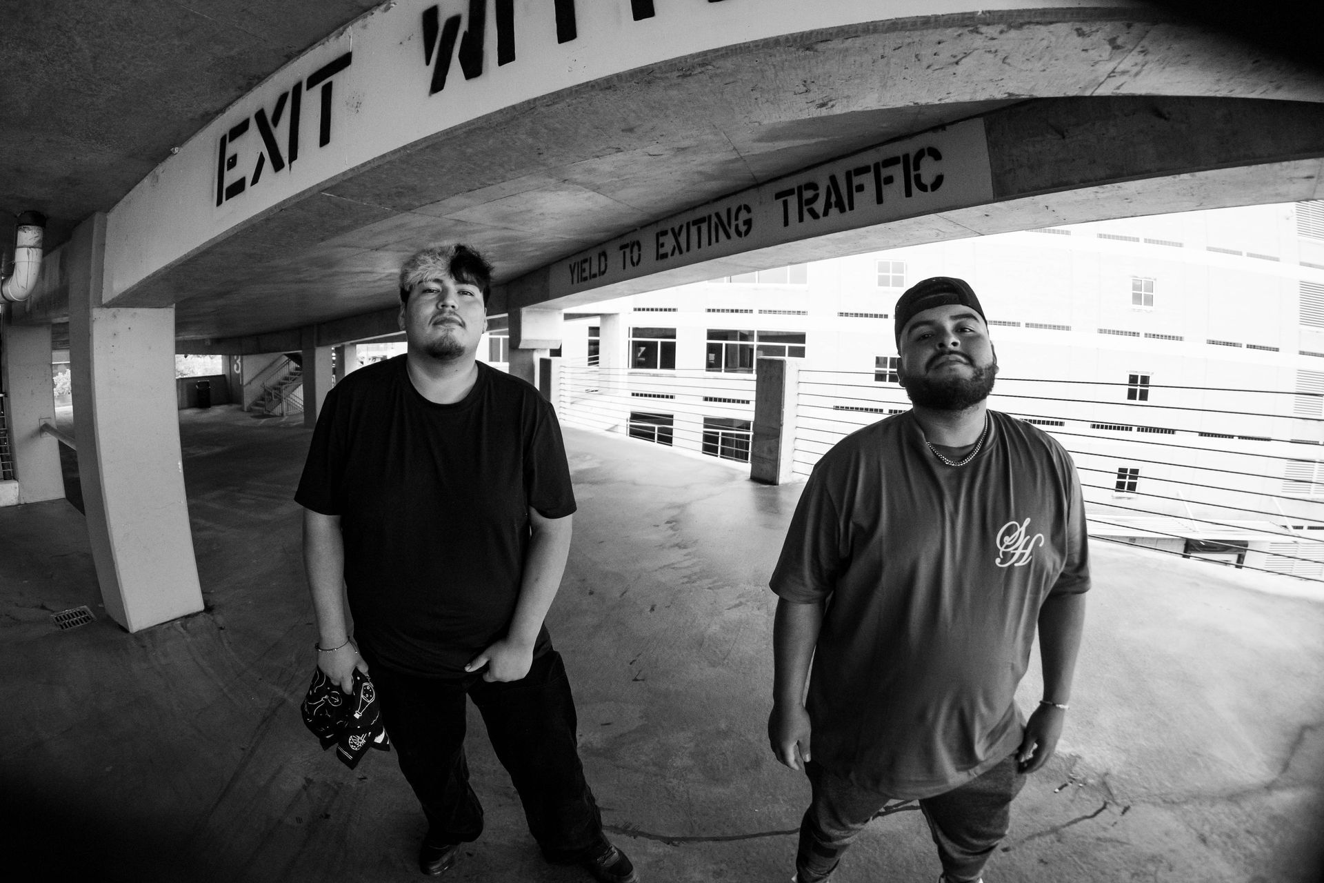 Two men standing under an exit sign in a parking garage, one holding a camera, in black and white.