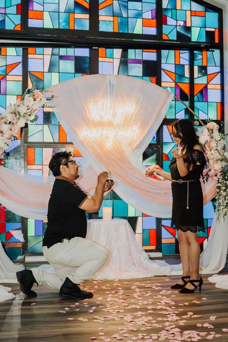 Man kneels proposing to a woman before a glowing draped arch in a stained-glass venue