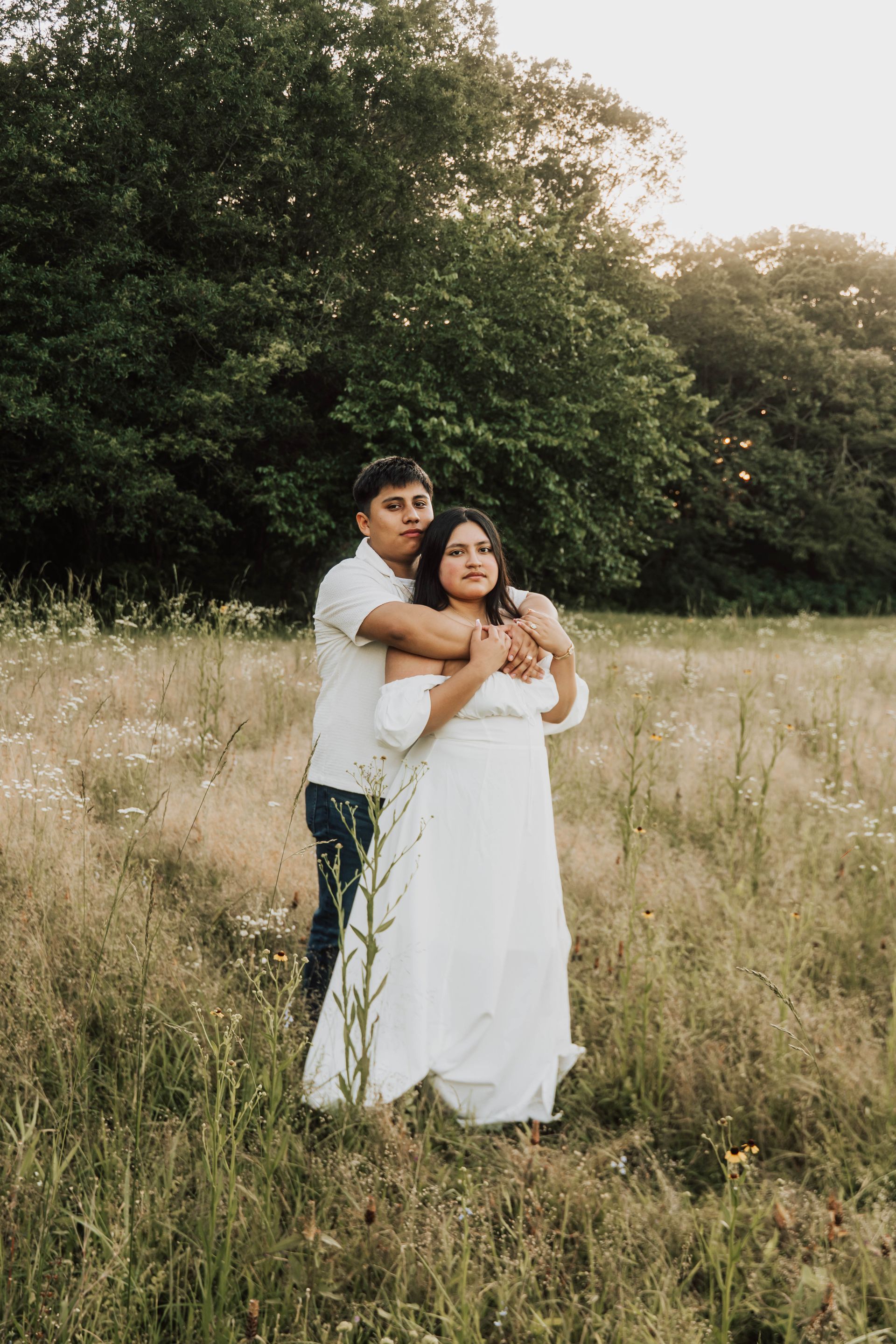 Couple embracing in a grassy field with tall trees in the background, the woman in a white dress