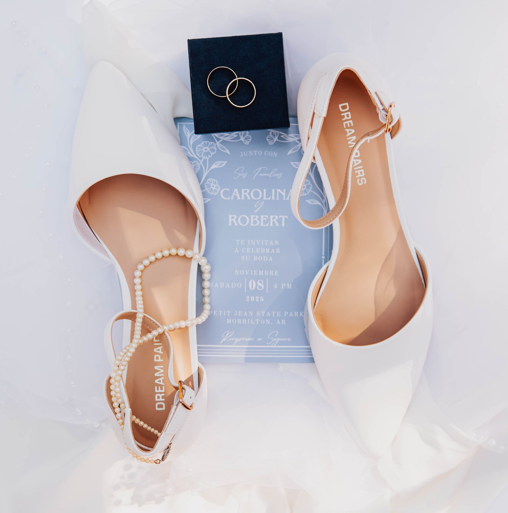 White bridal flats with pearls, blue invitation, and rings on a navy box on a white background