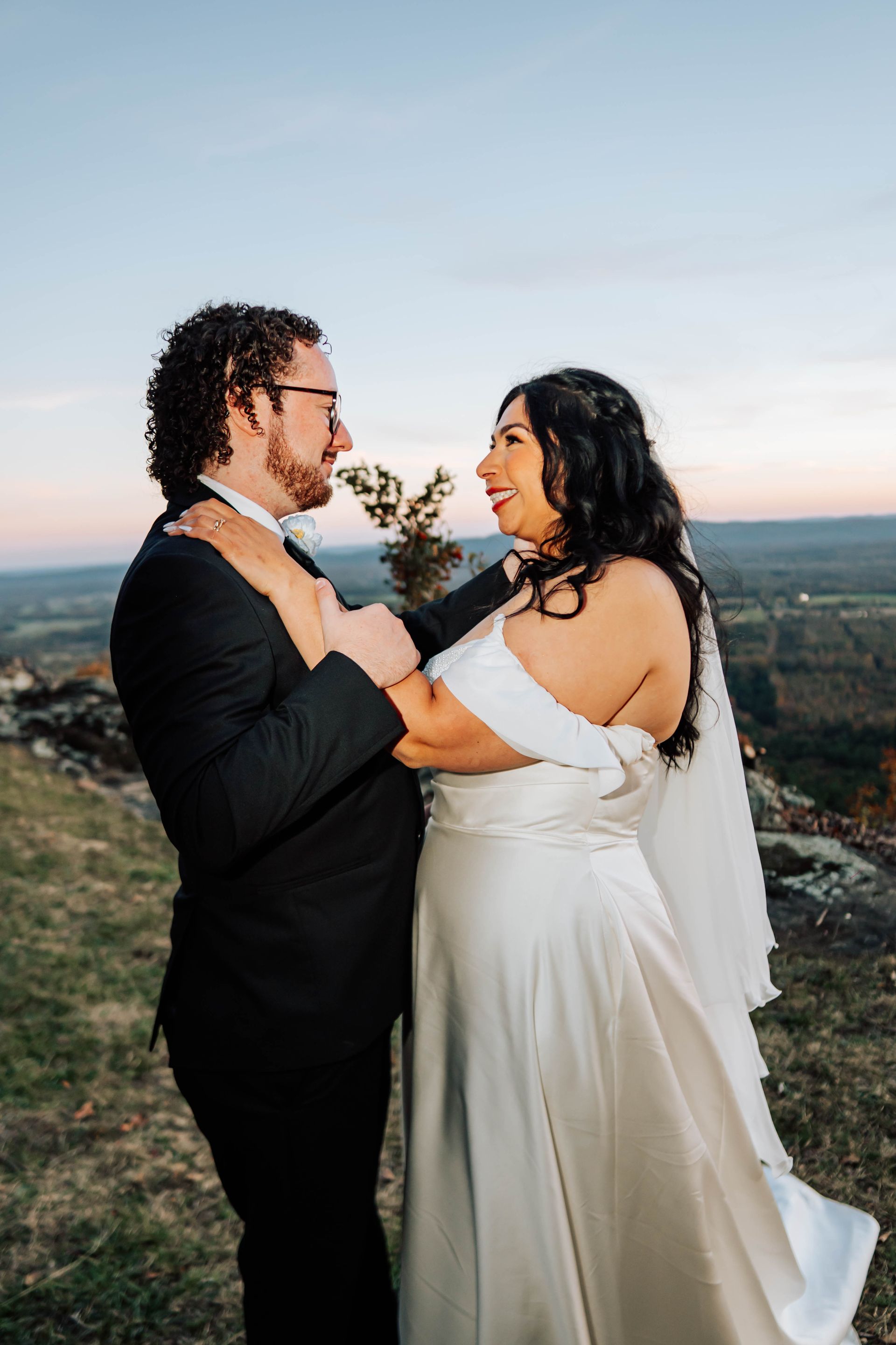 Two people in formal attire embrace outdoors at sunset, with a scenic landscape in the background.