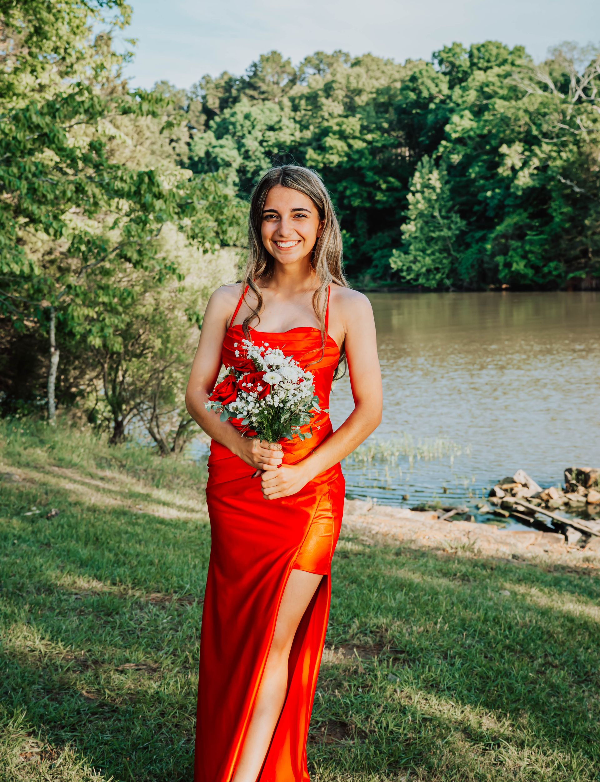 Woman in a red dress holding a bouquet by a riverside, with trees and water in the background.