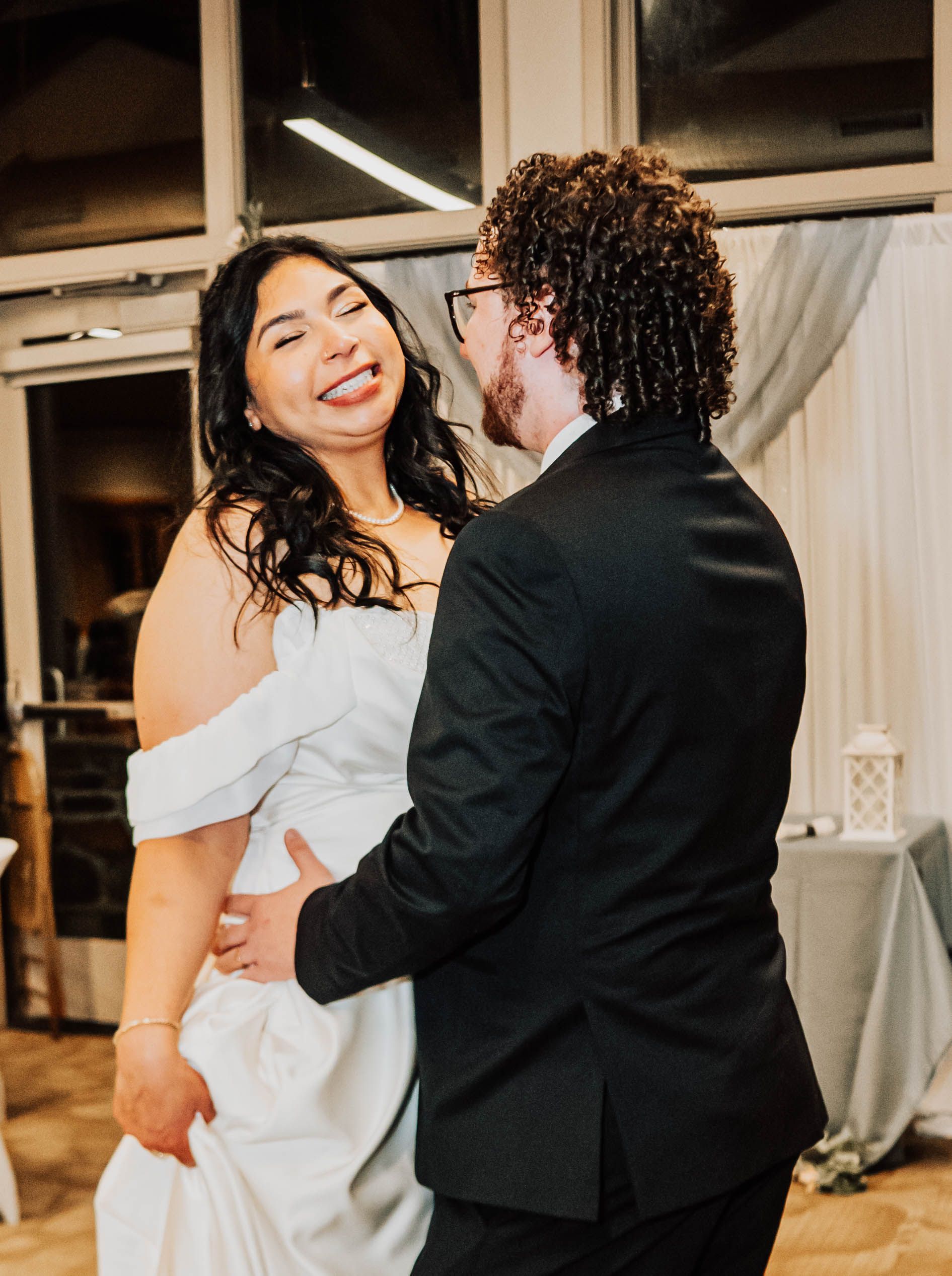 Couple in wedding attire sharing a dance indoors, smiling in a warmly lit room.