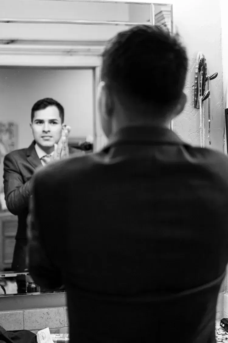 Man adjusting a tie in a mirror, with his back to the camera in a dim room.