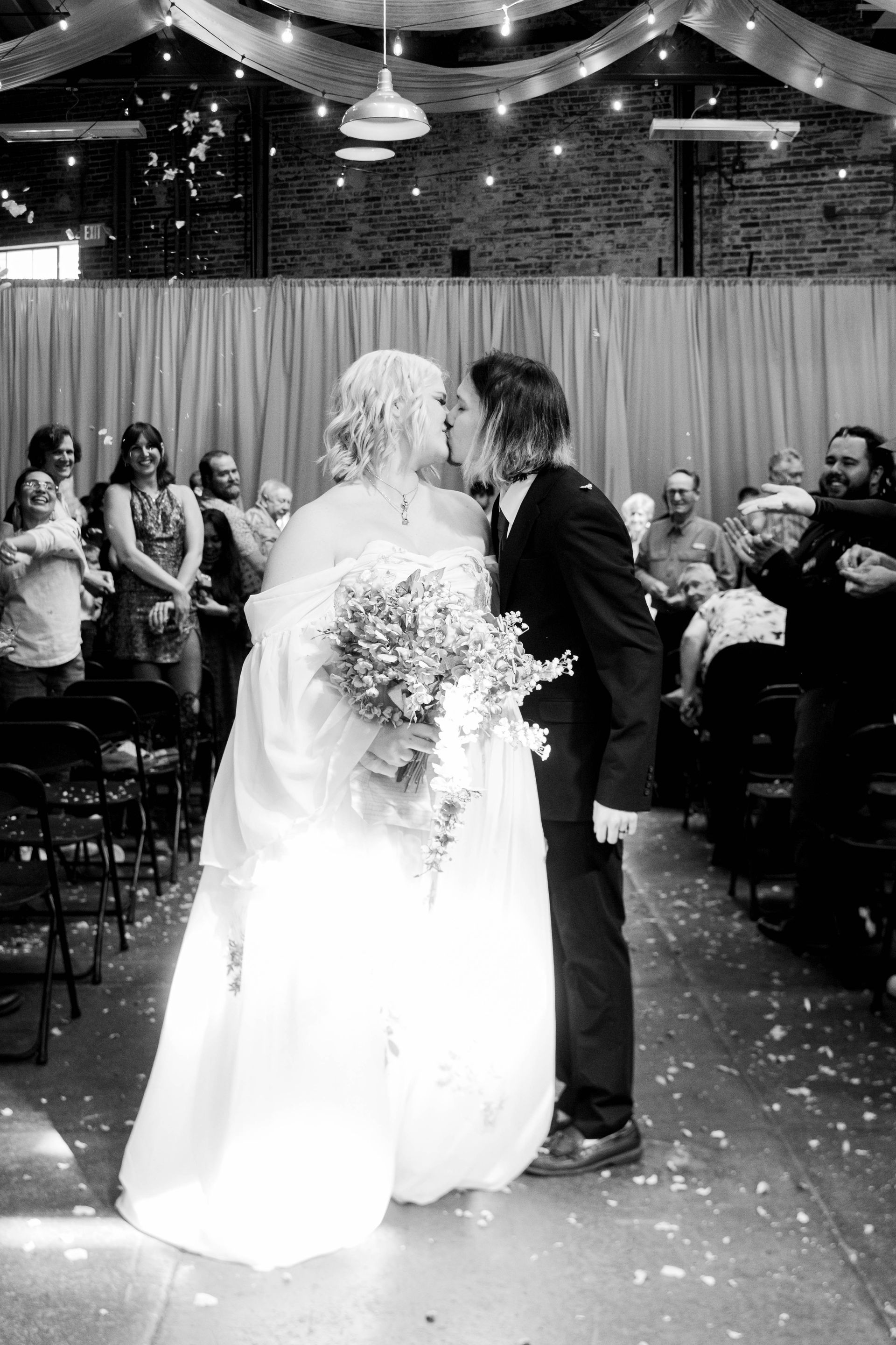 Bride and groom share a kiss at the altar during a wedding reception with seated guests and string lights.