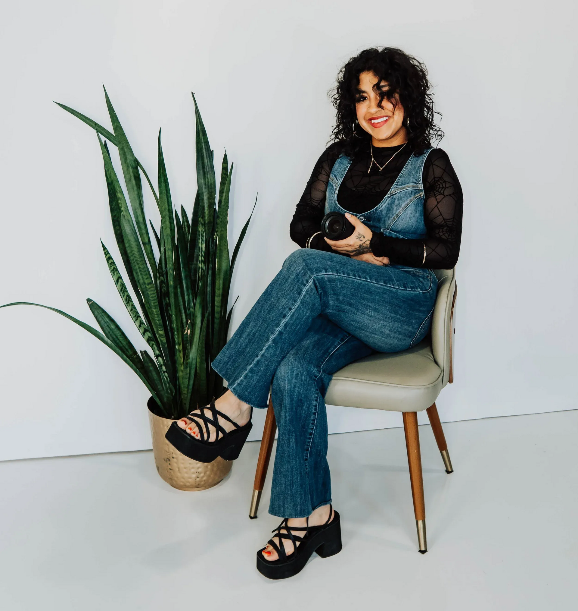 Woman seated in a chair beside a tall potted plant, wearing black top, blue jeans, and black heels.