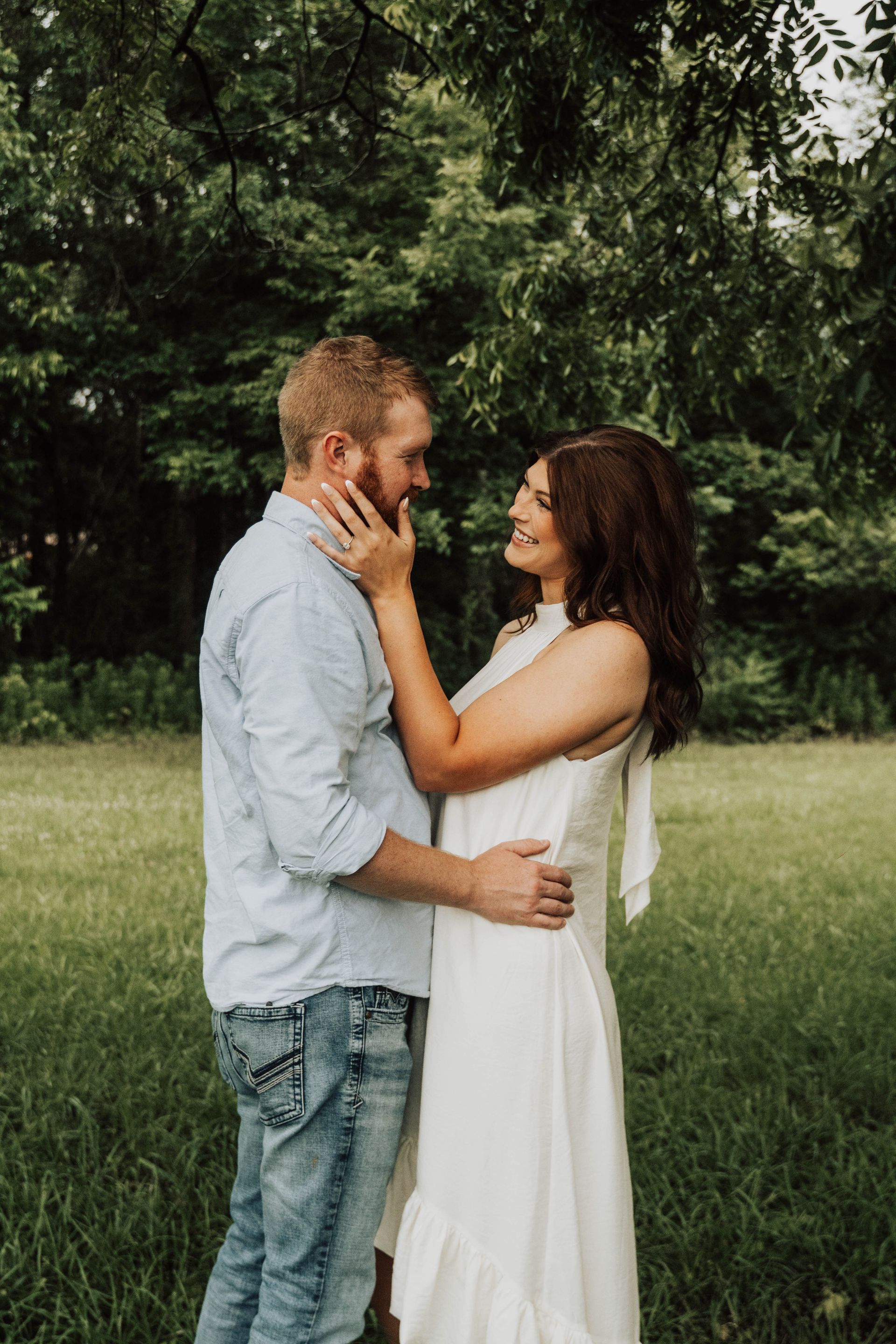 Couple embracing in a grassy park, standing close and smiling among trees.