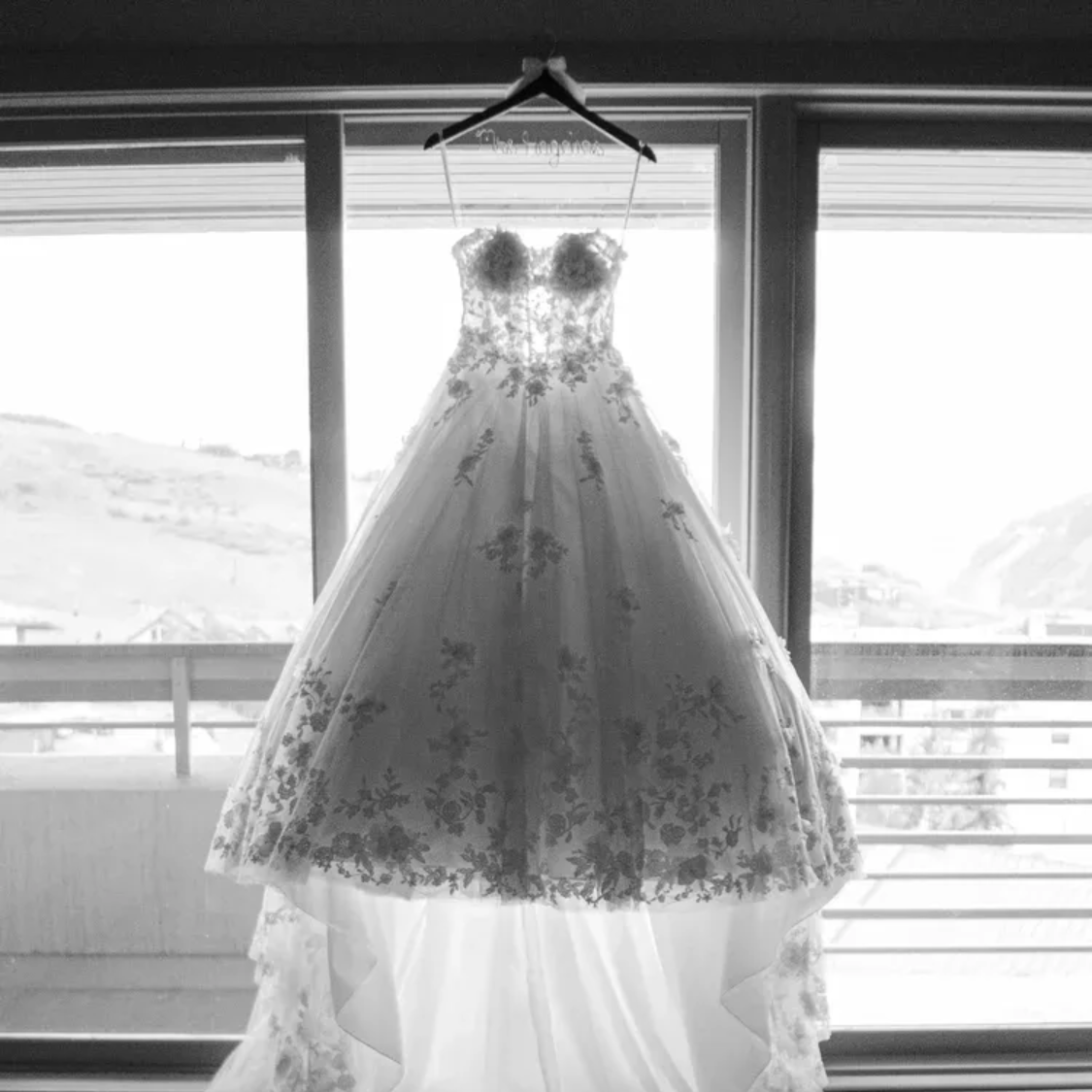 White lace wedding dress hanging in front of a window on a wooden hanger