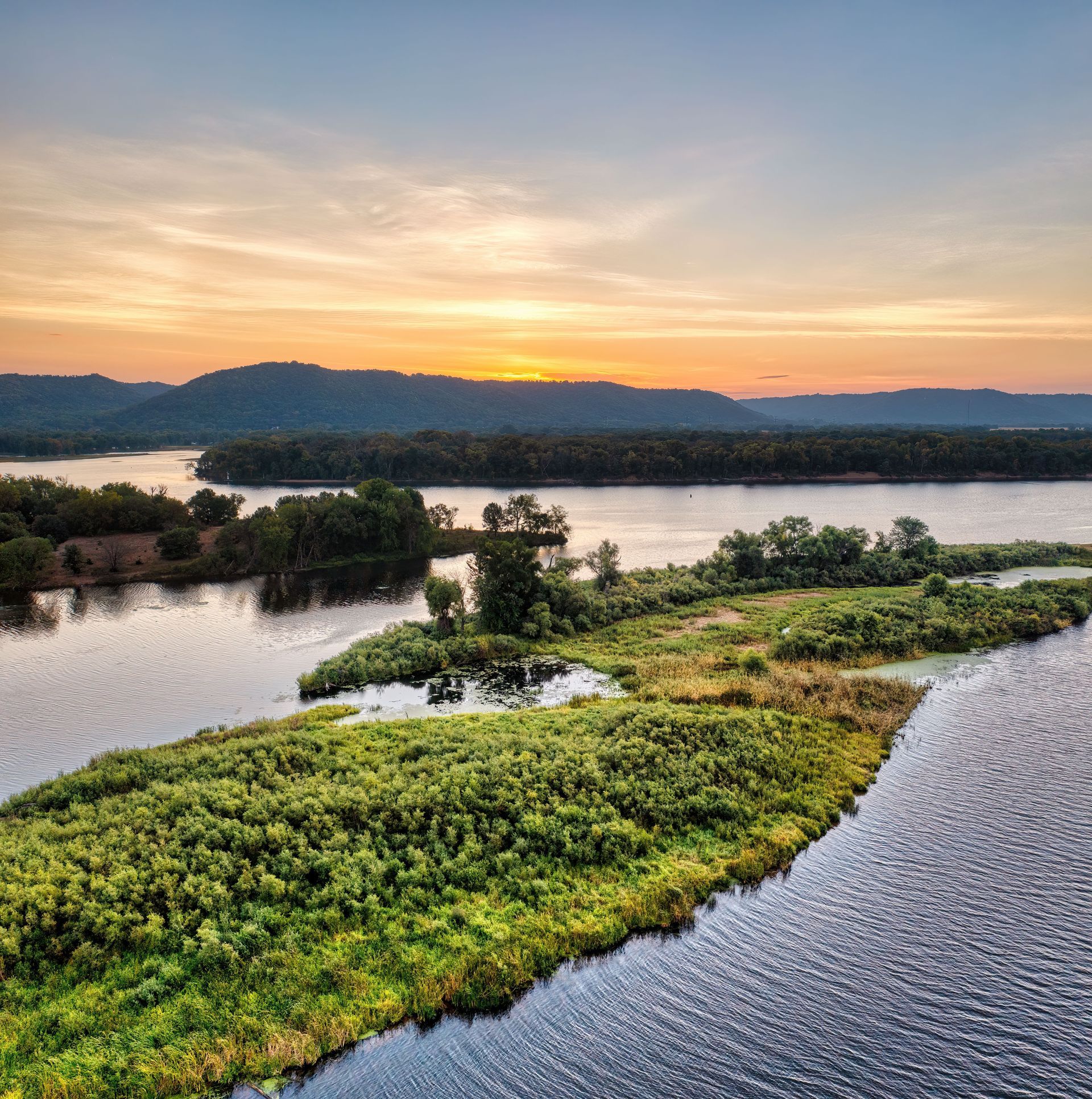 Sunset over a calm river with green islands and distant hills under a warm orange sky