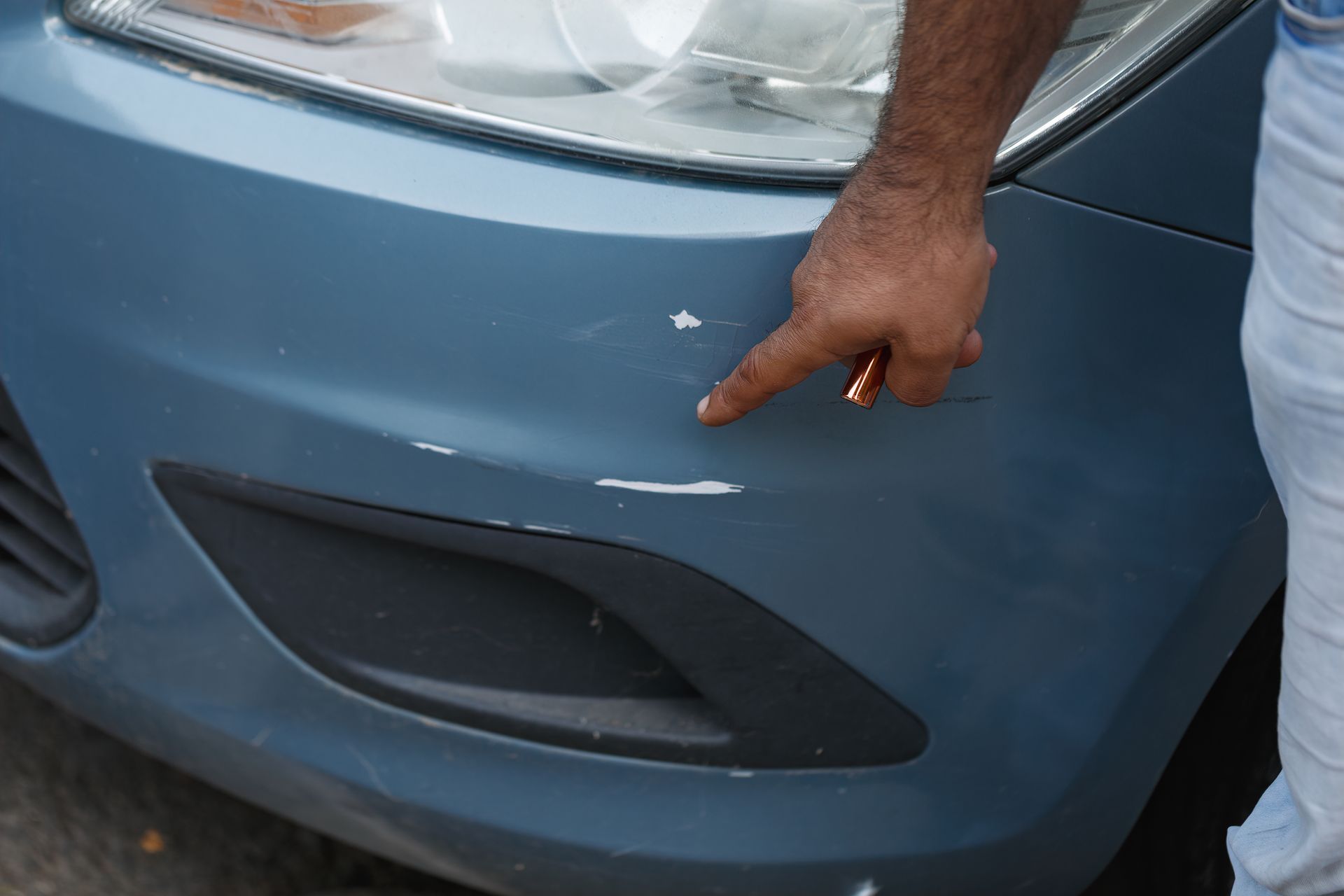 Hand pointing to a scratch on a blue car’s front bumper