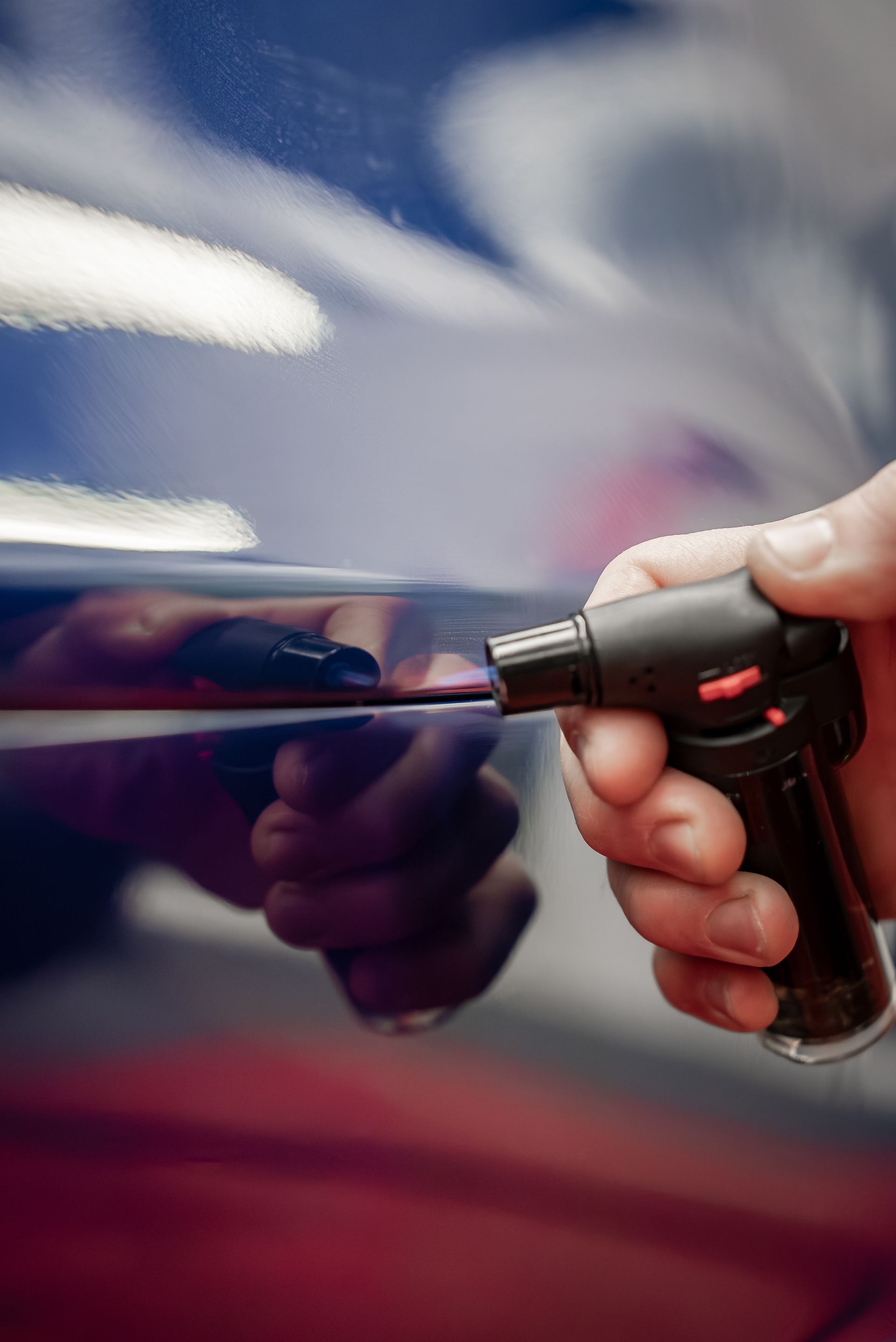 Hand holding a spray nozzle aimed at a shiny blue car panel, with a reflected arm visible.