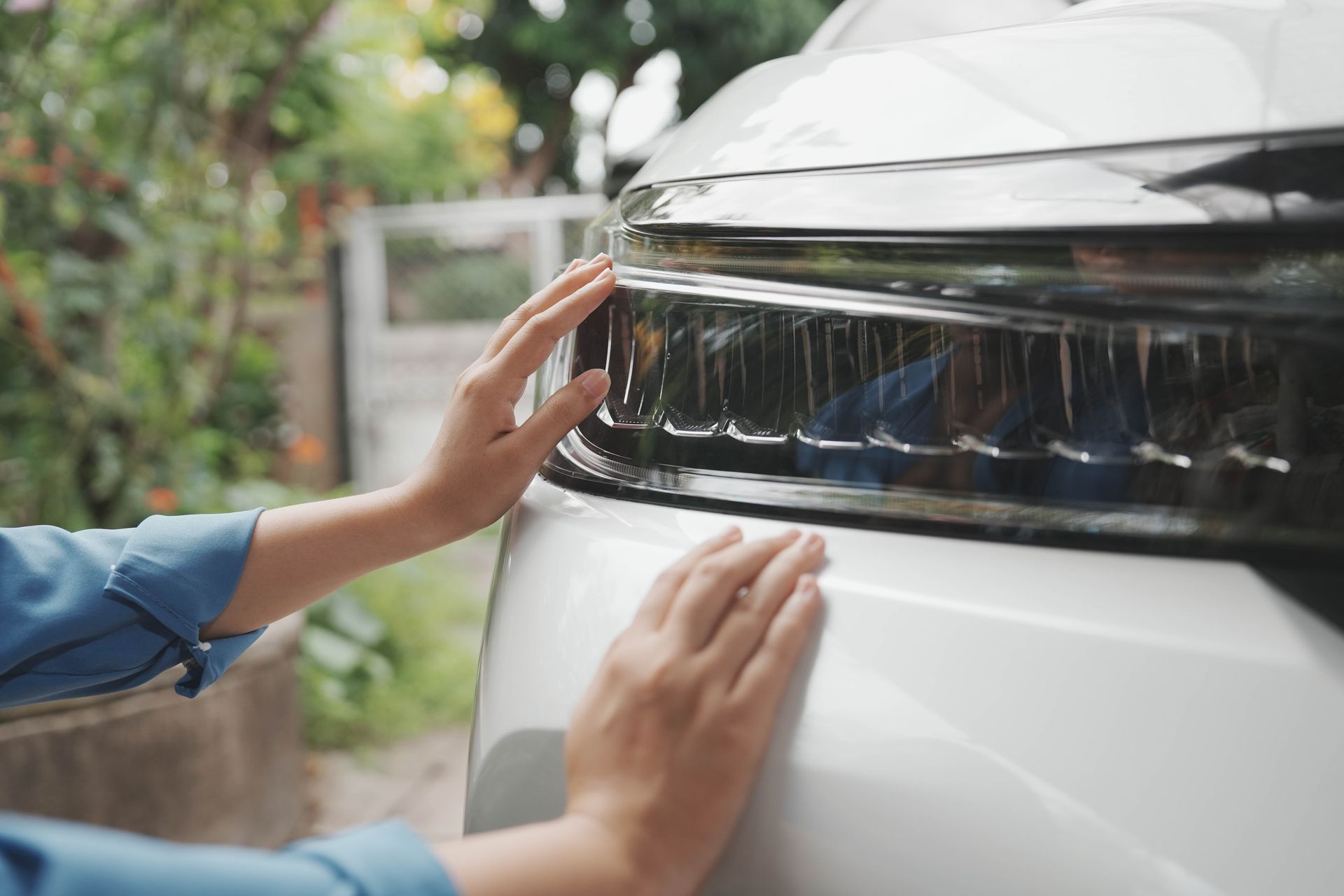 Hands polishing the front headlight and hood of a white car outdoors