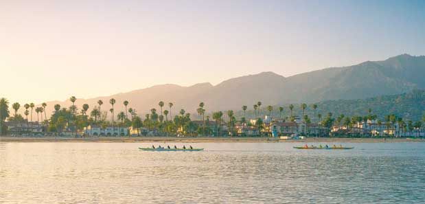 Calm shoreline with a small boat, palm trees, and mountains under a warm pastel sky
