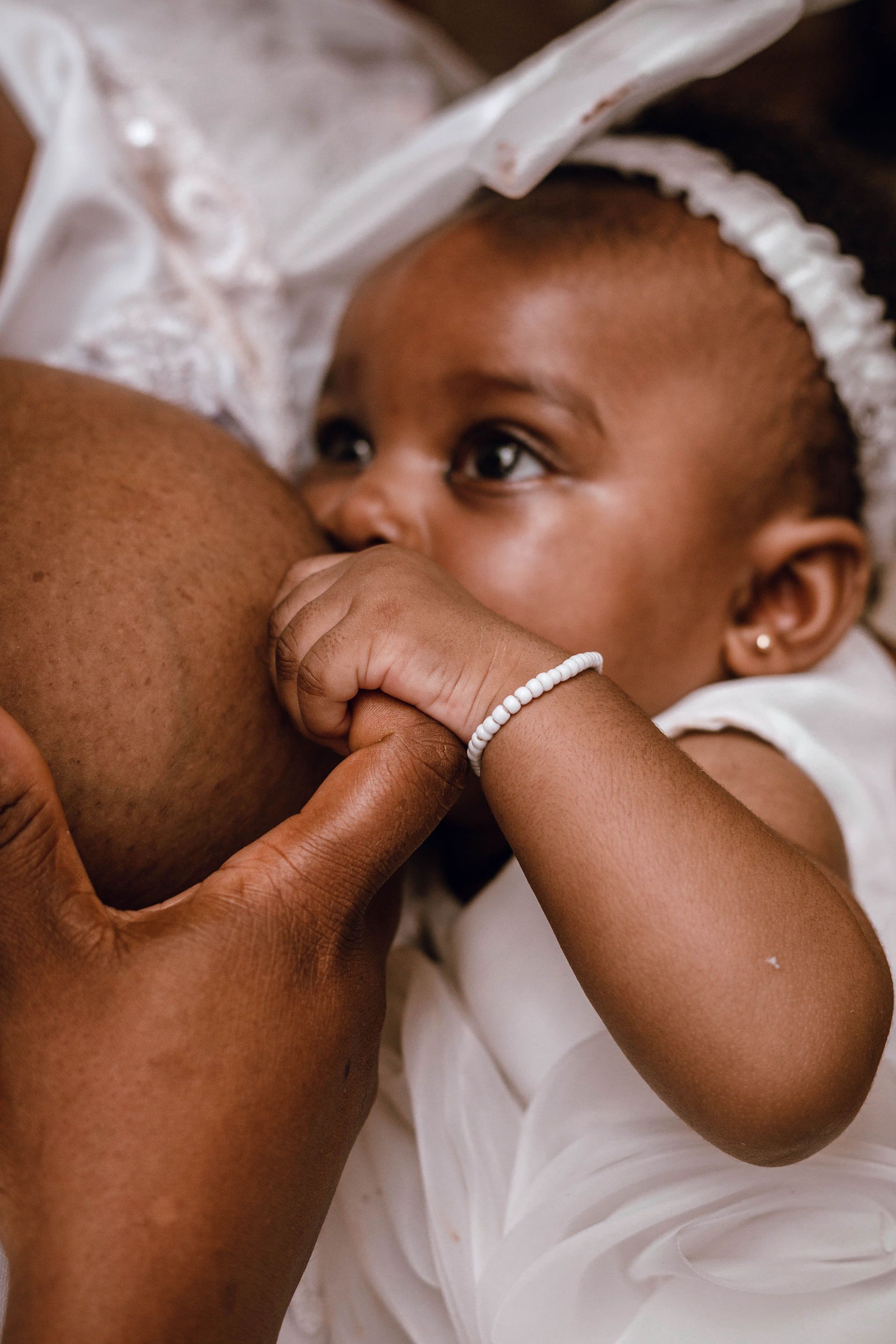 Baby in white dress breastfeeding, holding a hand and looking up attentively.