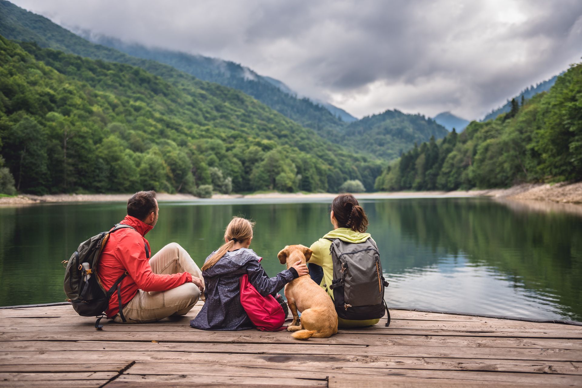 Four hikers with backpacks sit on a dock beside a calm lake surrounded by green mountains under cloudy skies