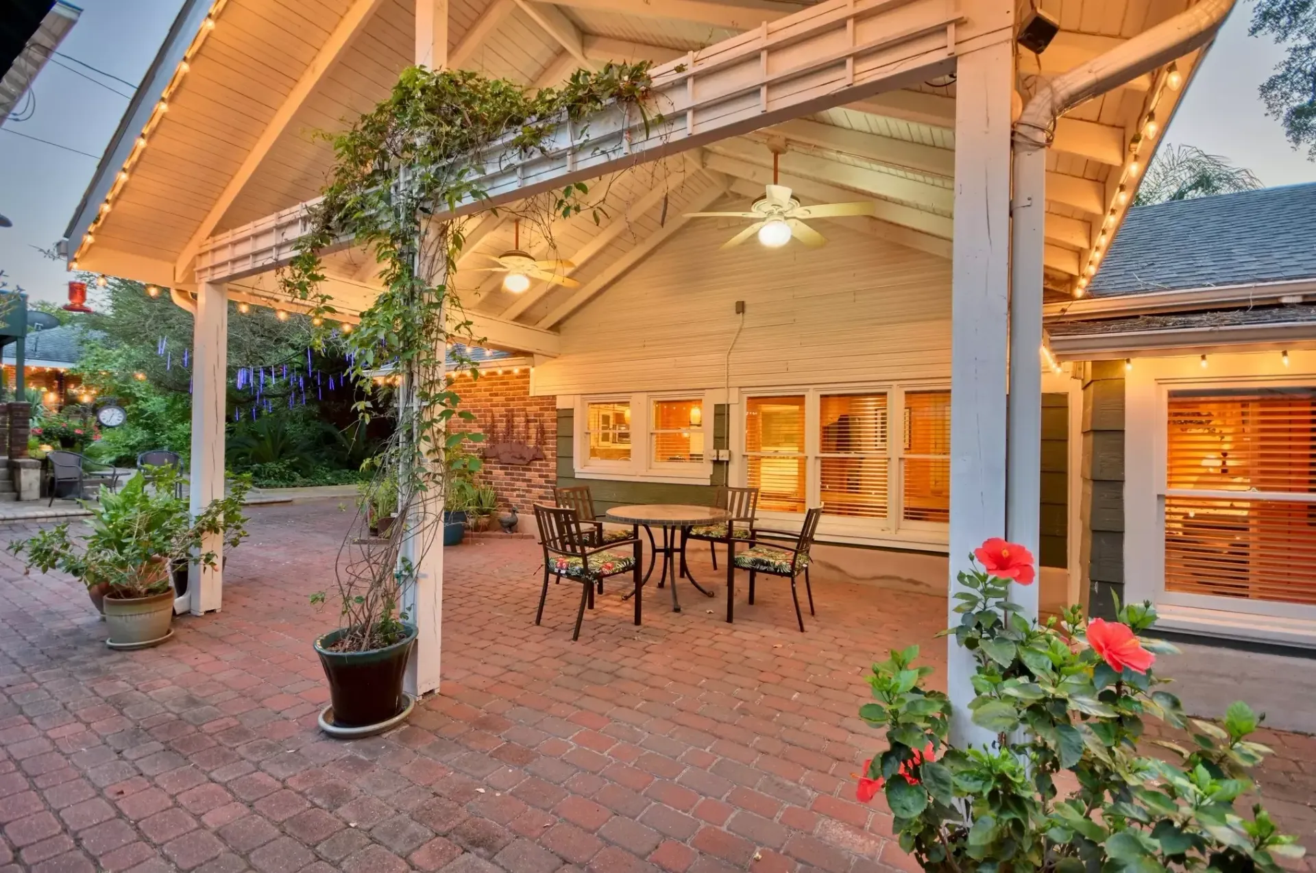 Covered patio with brick flooring, dining table, and flowering plants.