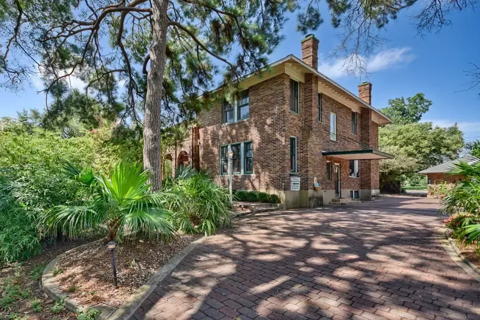 Brick house with a long driveway and lush landscaping under a tree.