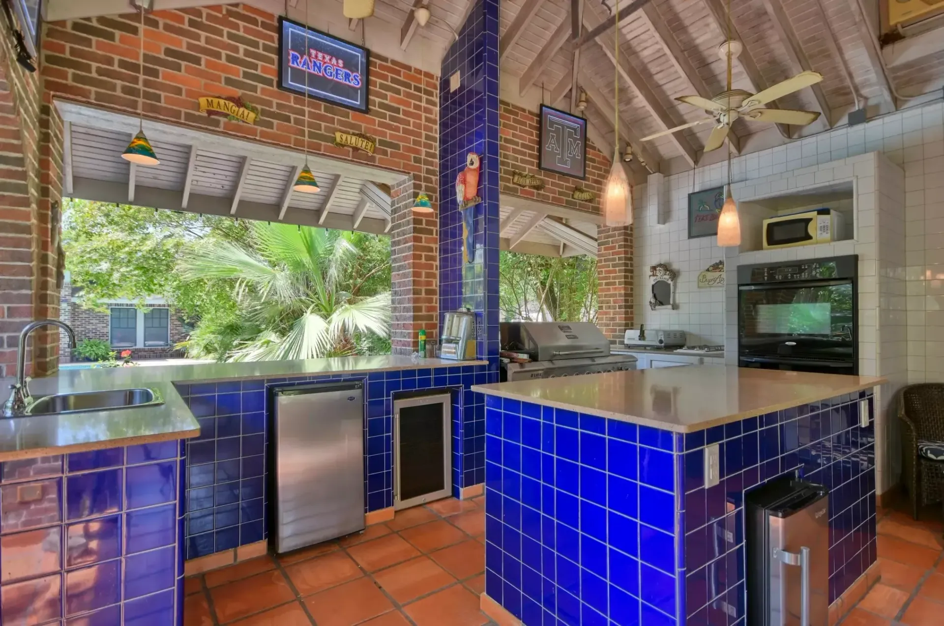 Outdoor kitchen with blue tile, brick, stainless steel appliances, and a view of trees.
