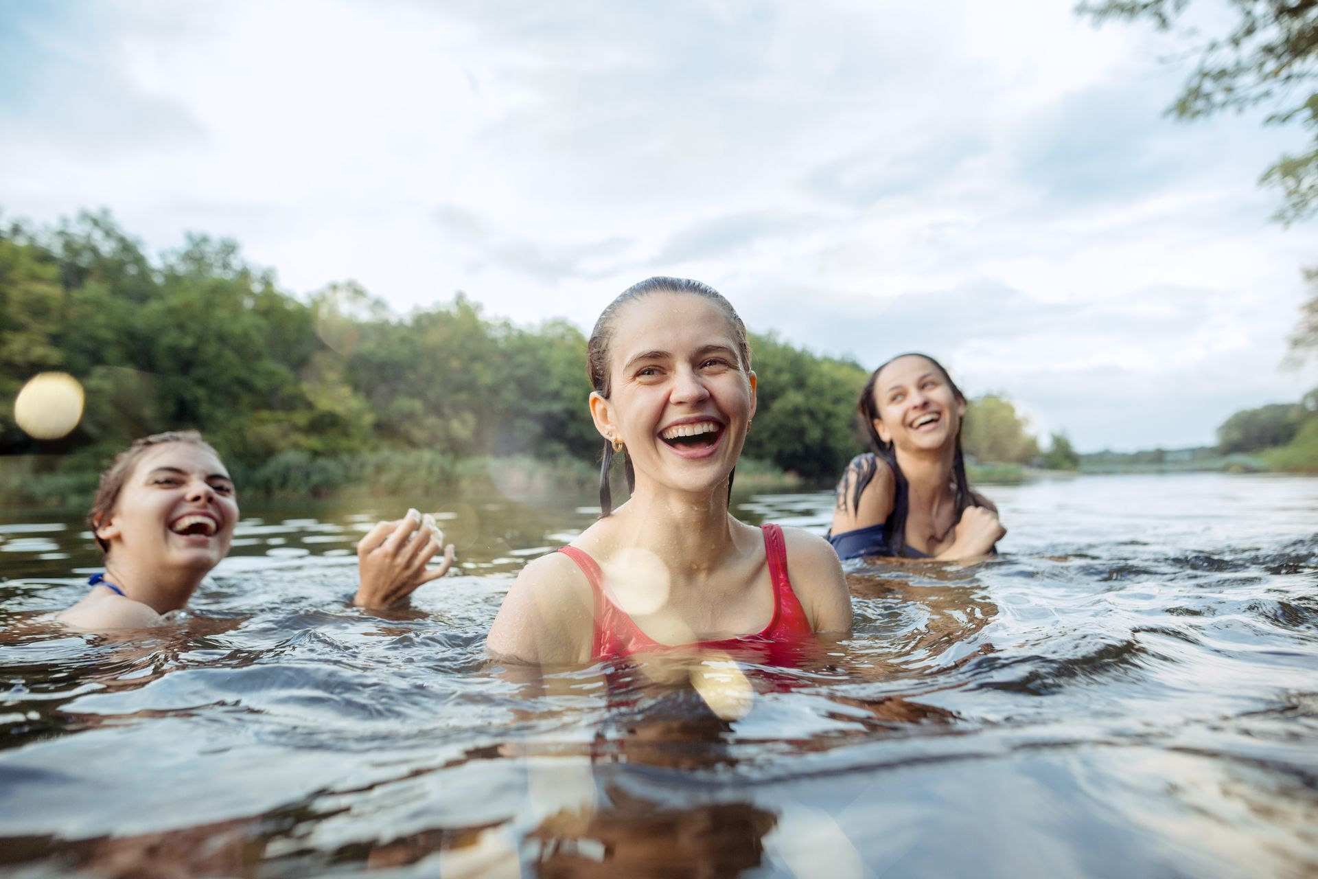 Three friends smiling and swimming in a river, with trees and cloudy sky in the background