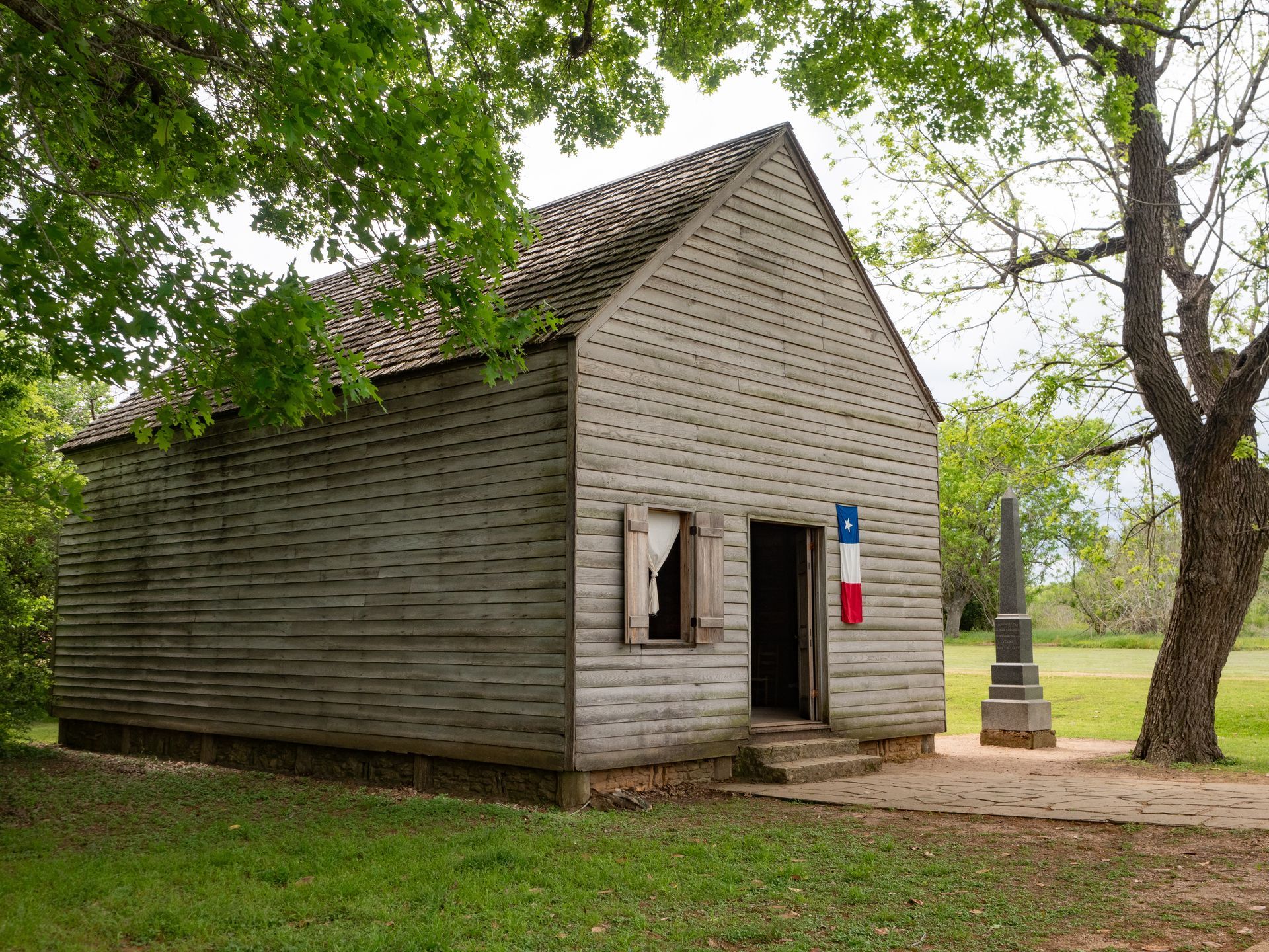 Small wooden cabin with shuttered windows in a grassy park under trees.