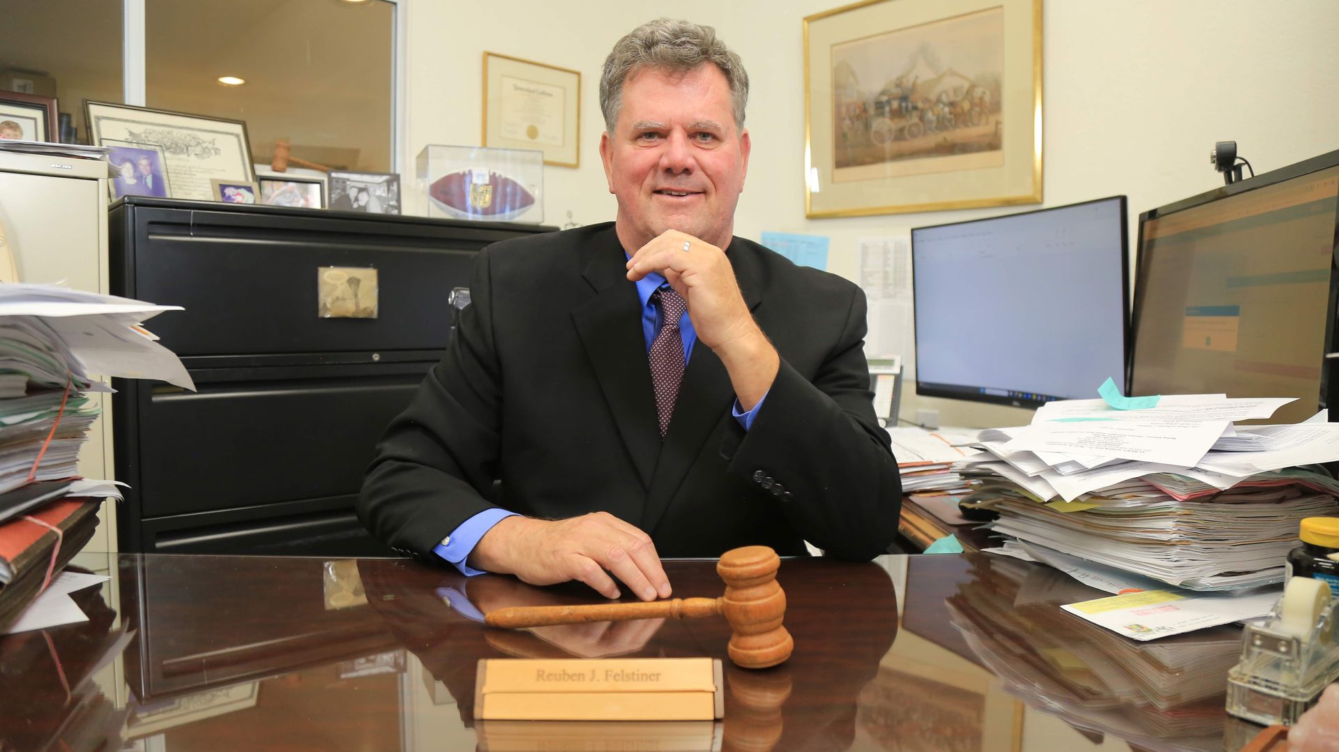 Man in suit seated at desk, holding gavel. Office setting, smiling, papers visible.