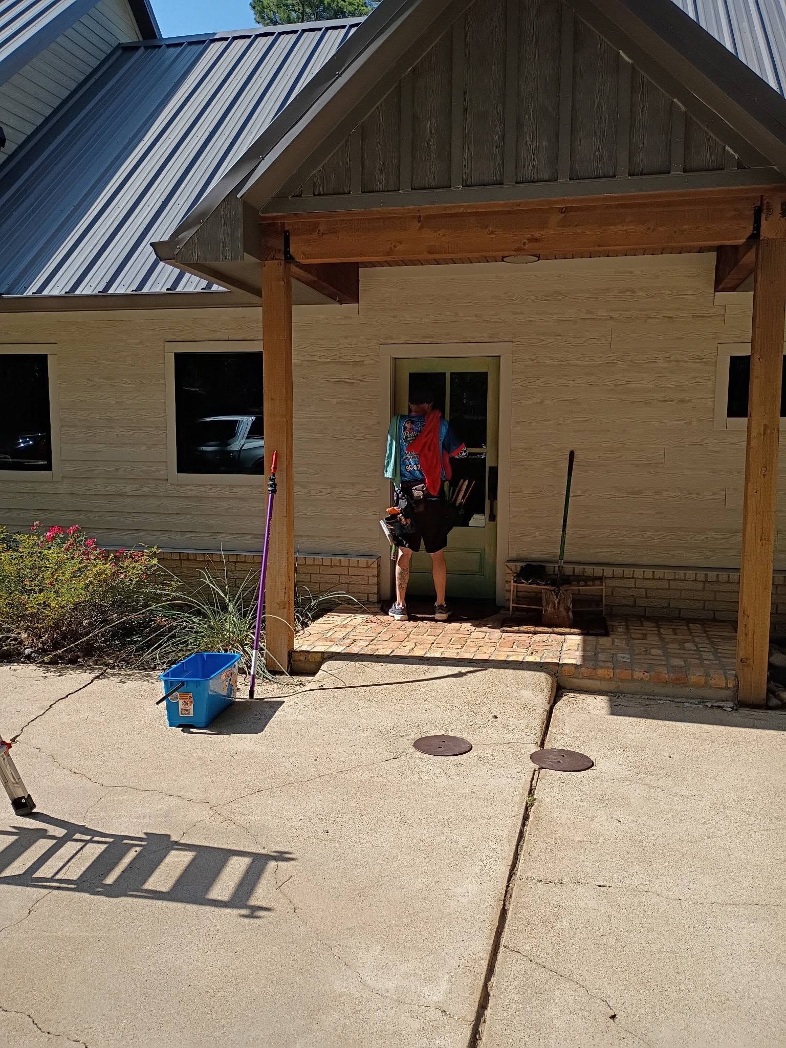 Person standing at a brick house doorway under a wooden porch, with a blue bucket on the concrete driveway.