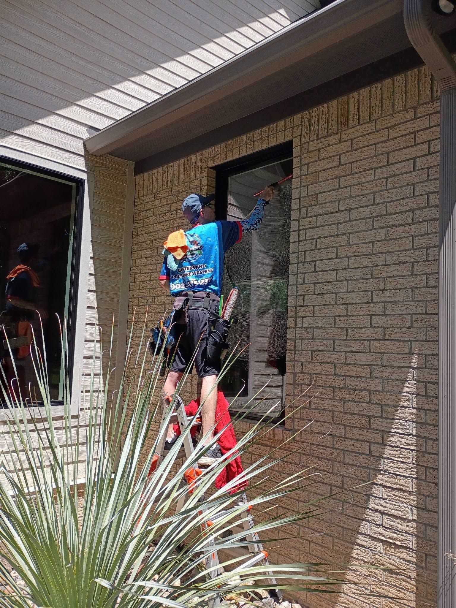 Person in blue shirt stands on a red ladder, painting a wall beside a brick house.