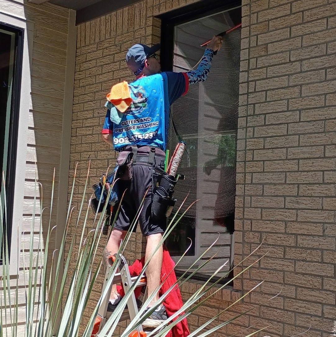 Worker spray-painting a brick house exterior beside a doorway, with a ladder in the foreground