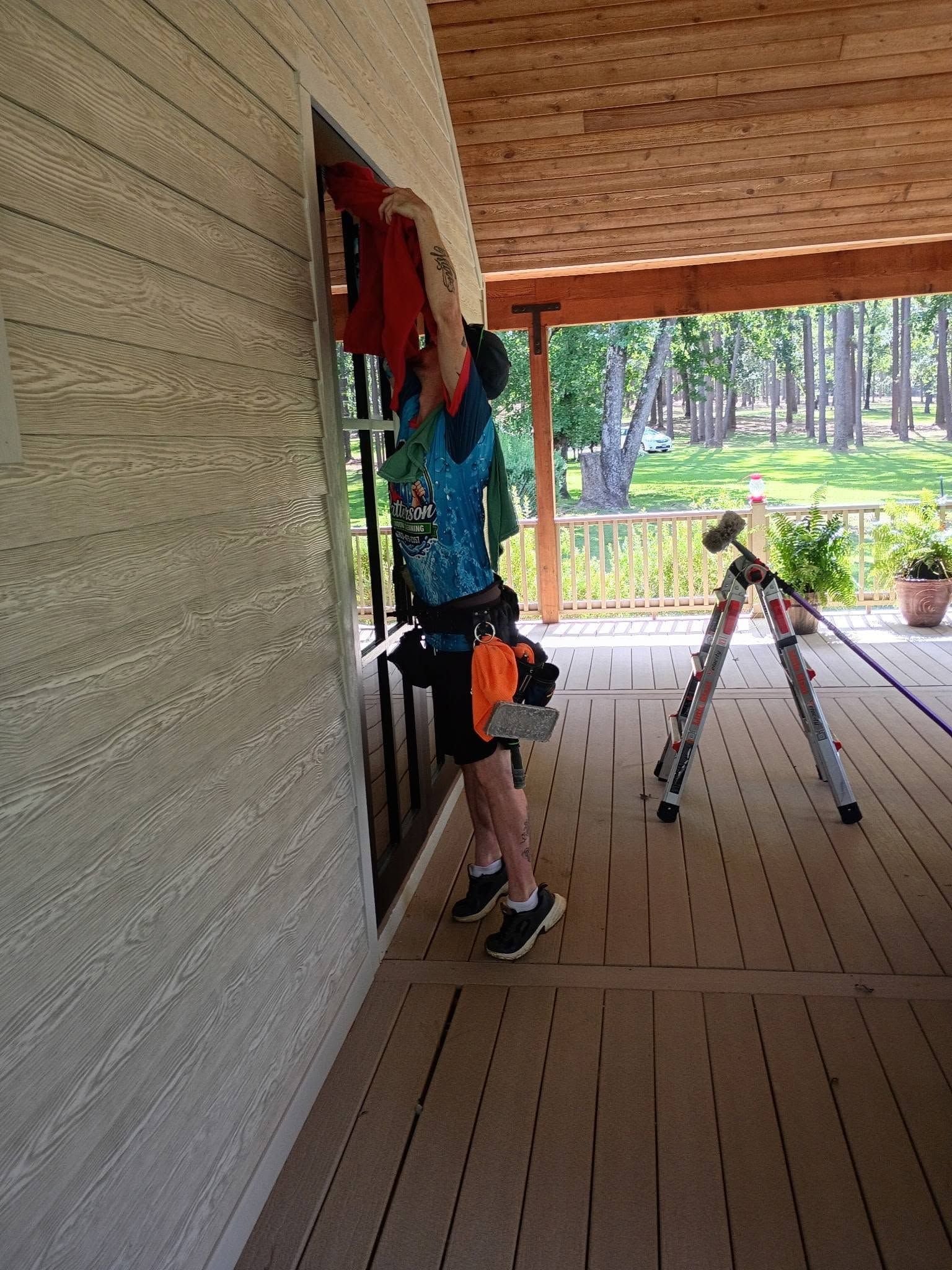 Person on a porch adjusting a red wreath by a wall, with a tripod nearby and trees in the background.