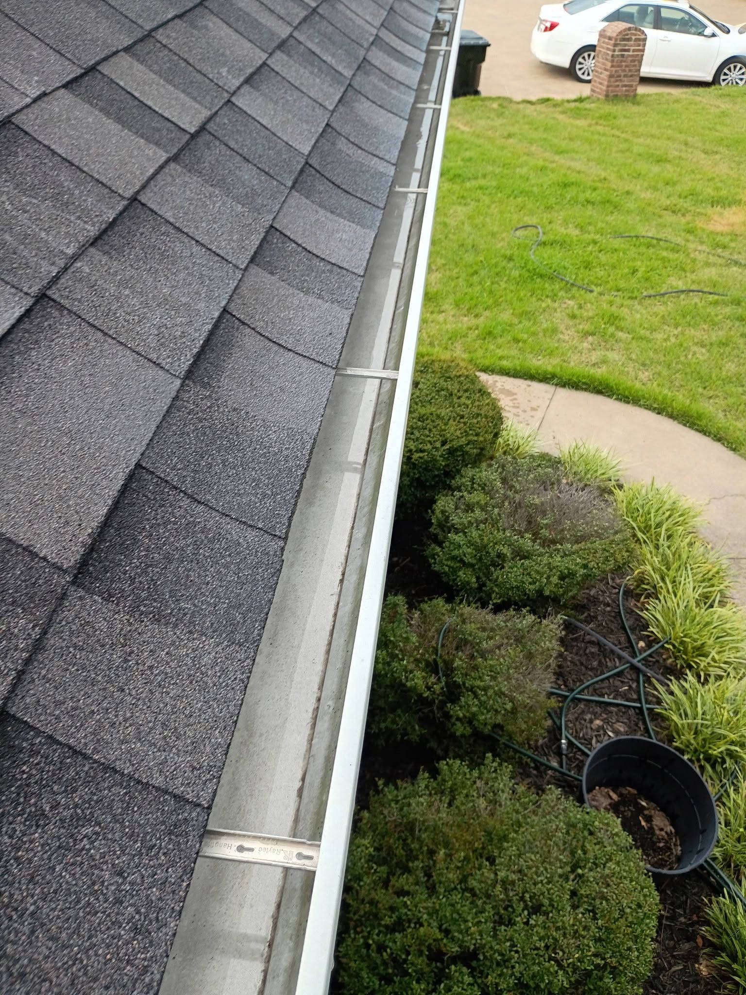 Roof edge and gutter beside a manicured lawn and shrubs, viewed from above.