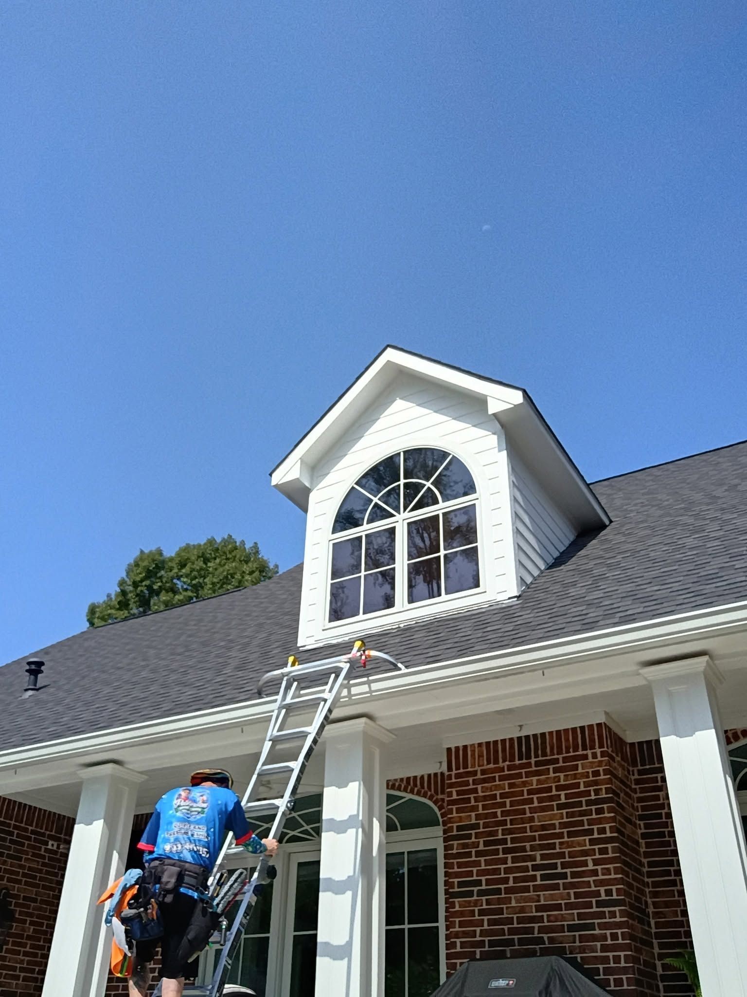 Worker on a ladder cleaning a dormer window on a brick house under a blue sky