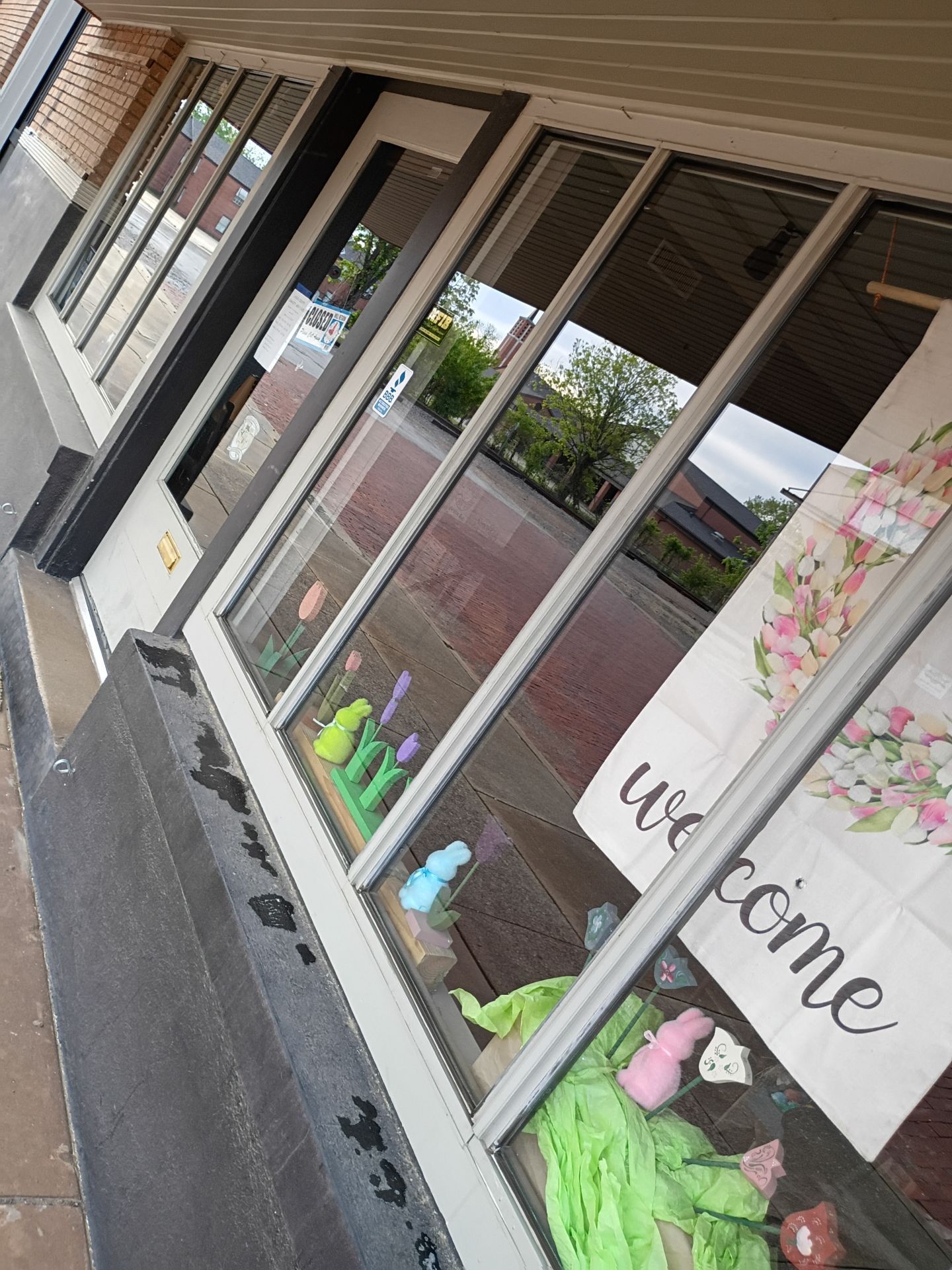 Storefront windows with “welcome” sign and potted flowers along a sidewalk
