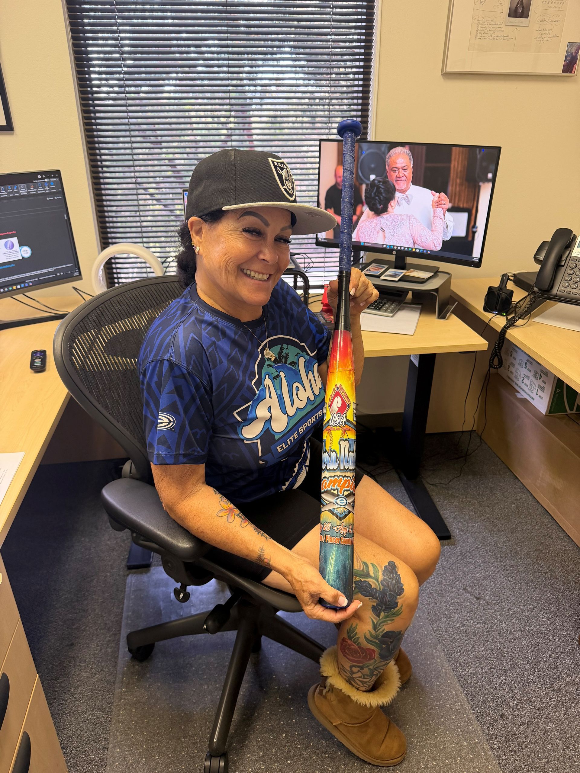 Person smiling in an office, holding a colorful decorated object beside a desk with two monitors.