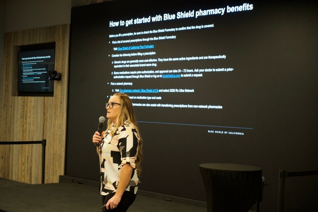 Presenter speaking at a conference beside a slide about getting better medication benefits, with a large screen behind her.