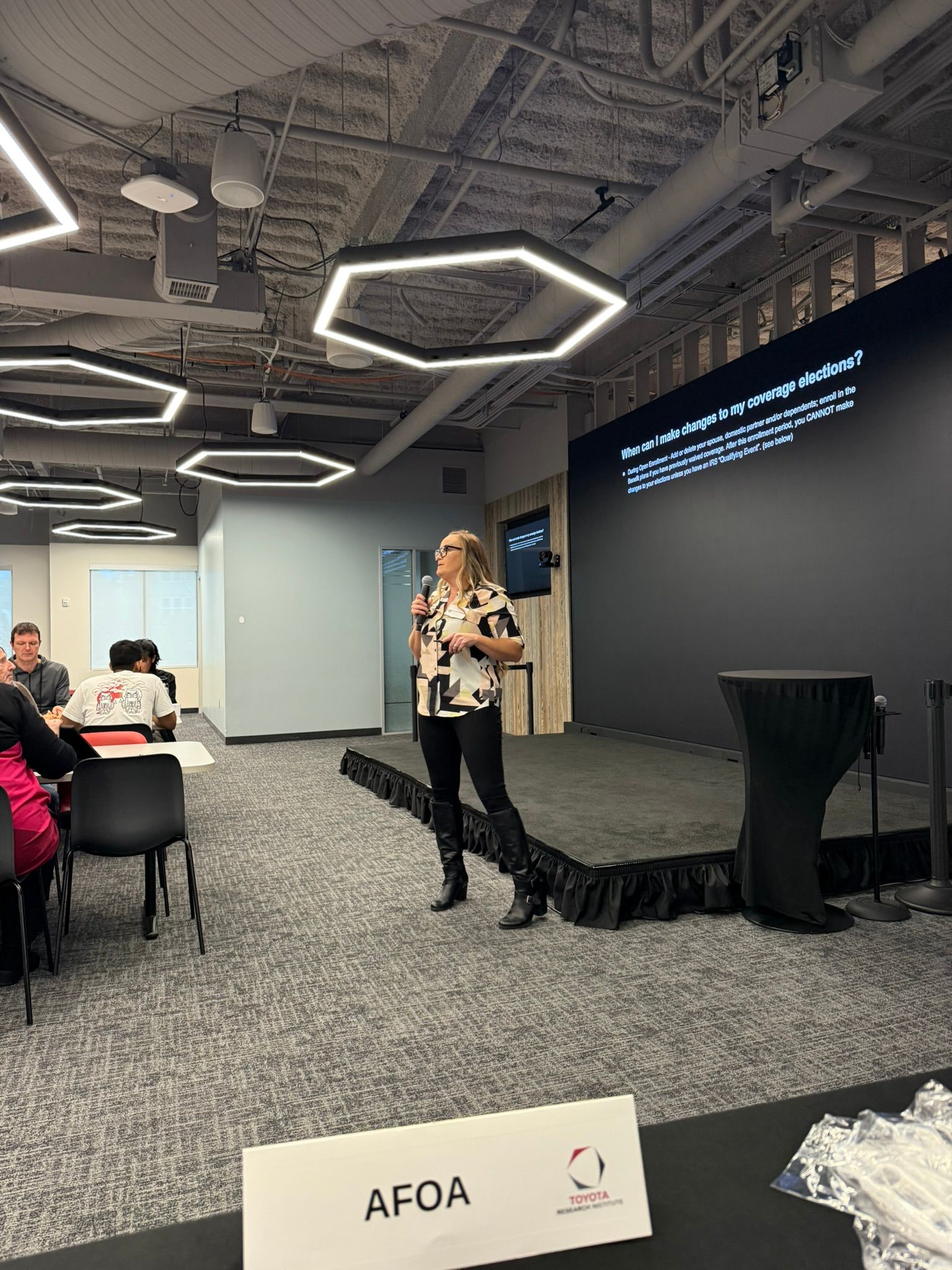 Speaker presenting on stage in a modern conference room, with seated audience and slide on dark screen.