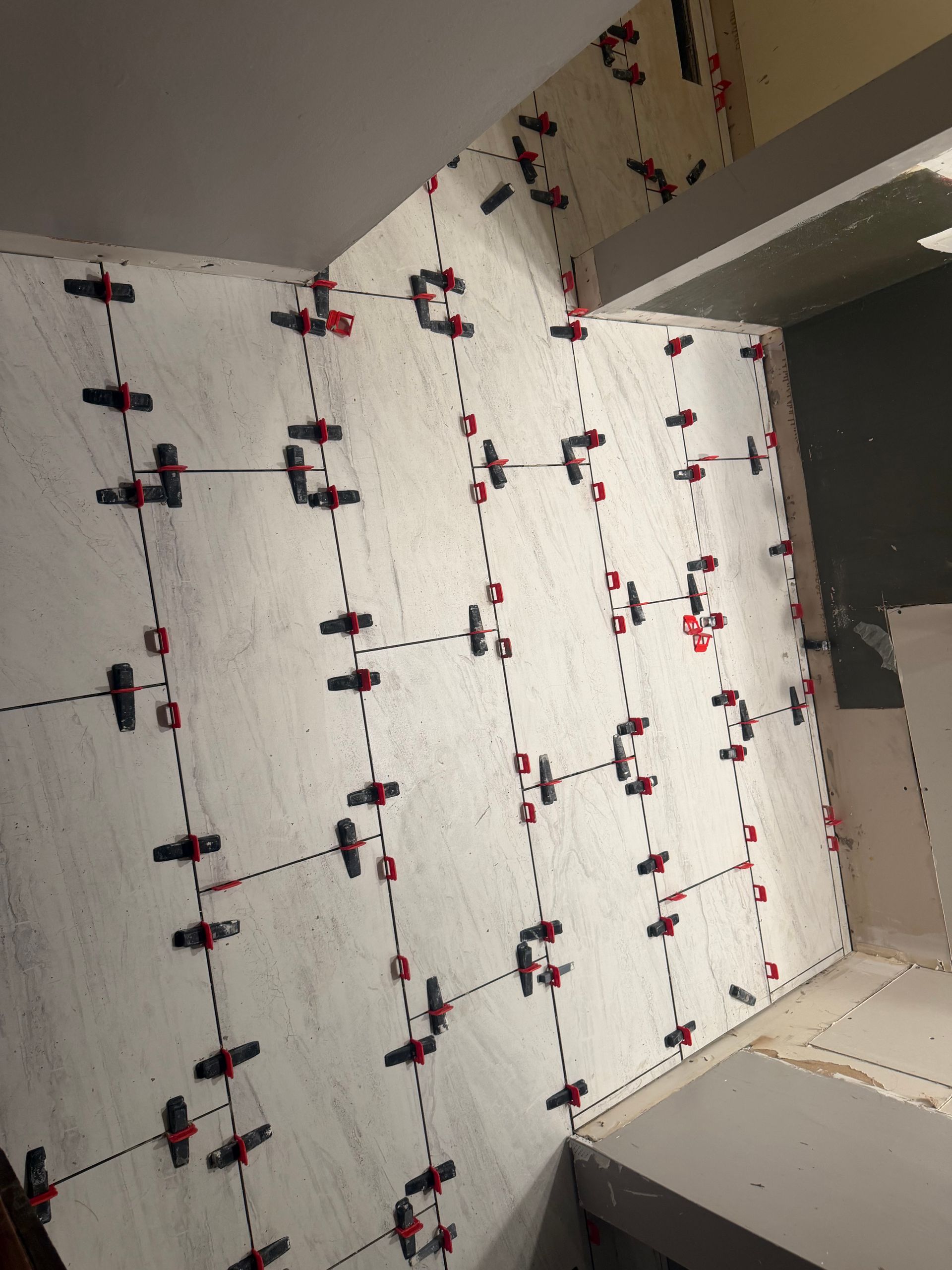 Tiled shower wall with white panels, black and red clips, and a doorway on the right.