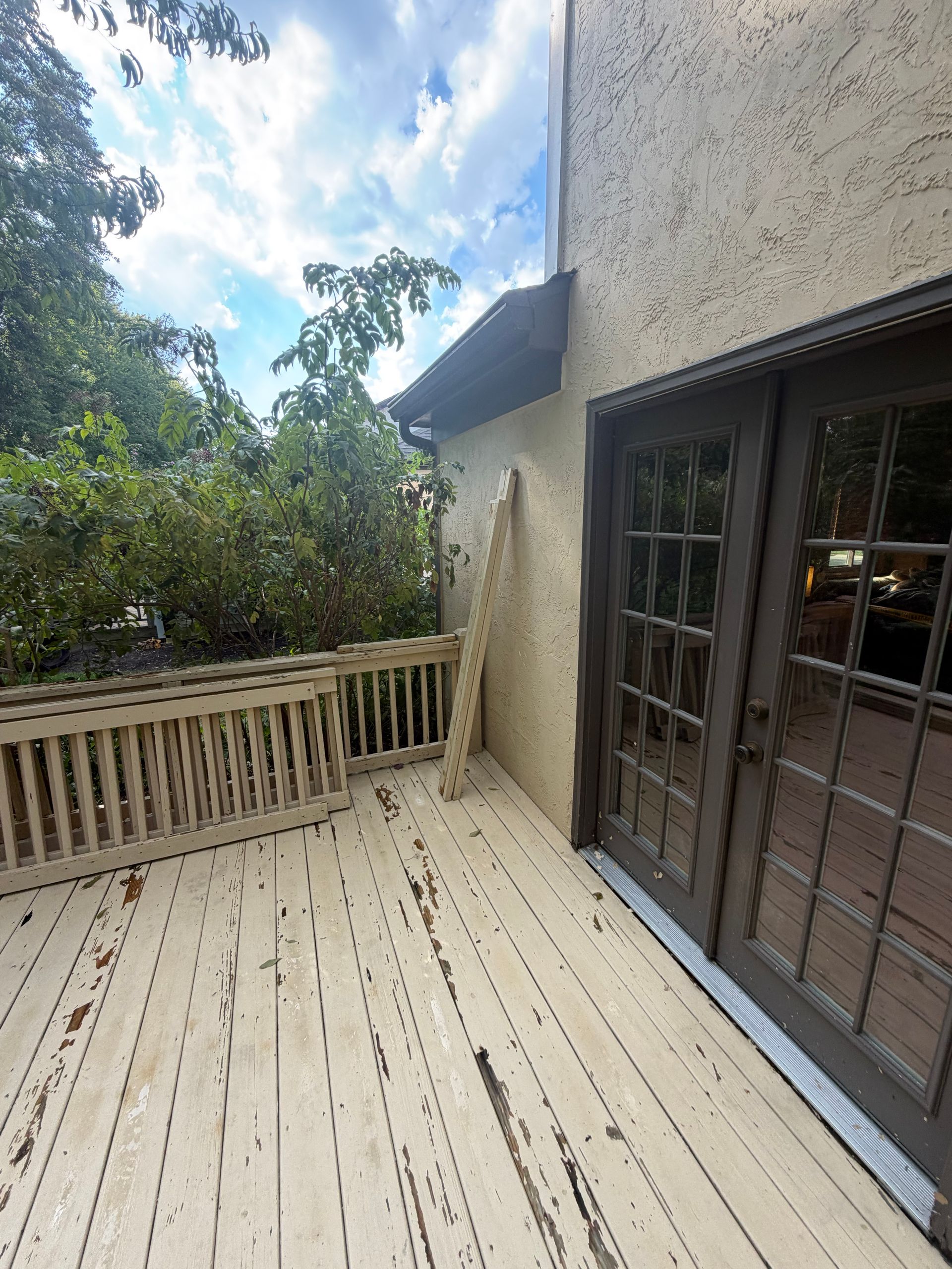 Wooden deck with railing beside a stucco house and glass patio doors, overlooking trees and sky