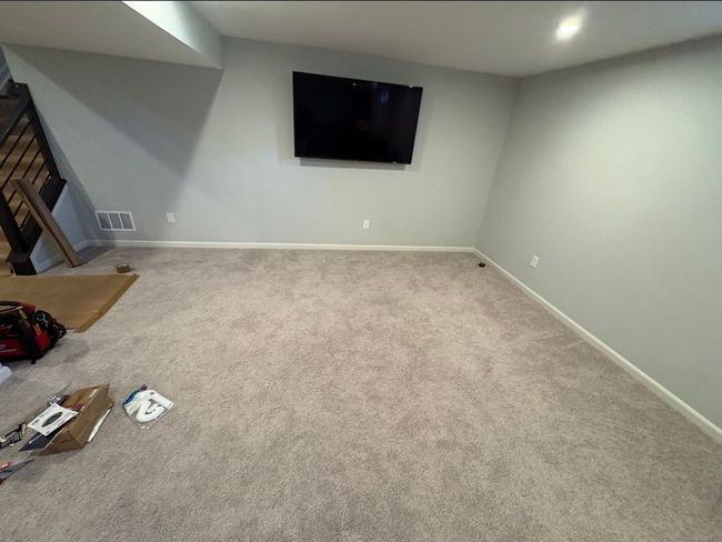 Empty carpeted living room with wall-mounted TV and stairs on the left