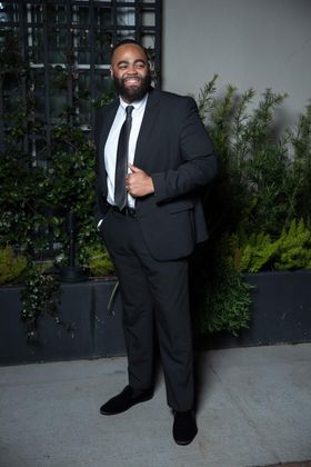 Man in a black suit adjusts his tie, standing outside.