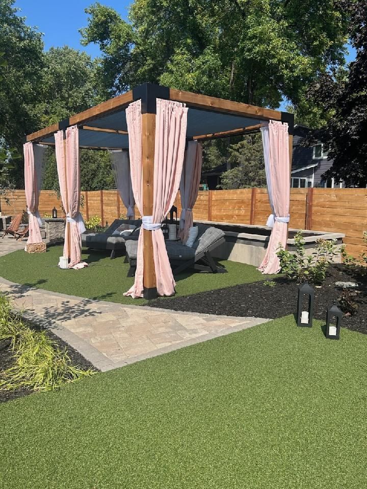 A wooden pergola with pale pink curtains stands on a grassy lawn with lounge chairs and a stone patio.